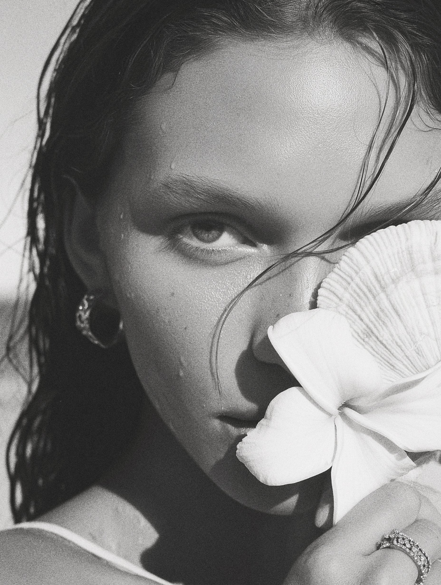 Close-up black and white photo of a woman holding a large flower near her face, covering her nose and mouth, with wet hair and a ring on her finger.