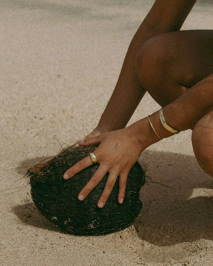 A person with jewelry on their hand and wrist is kneeling on sand, pressing their hand into a round, dark object resembling a coconut.