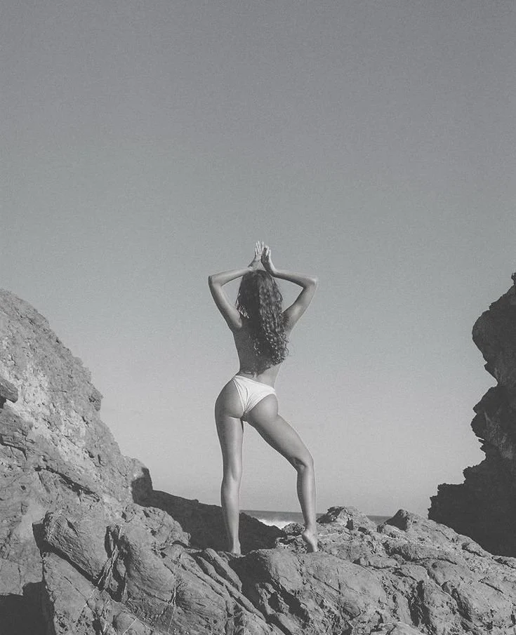 A woman in a swimsuit stands on rocky terrain, facing away, with arms raised and hands pressed together above her head, on a clear day at the beach.