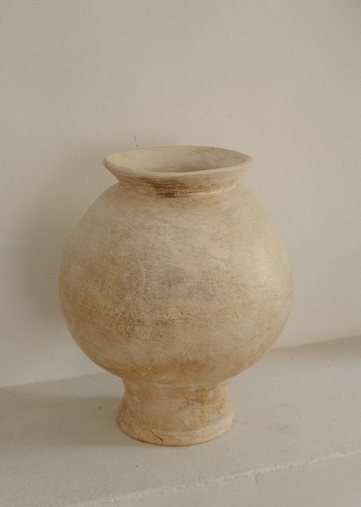 A cream-colored, round ceramic vase with a small, flared opening, positioned against a plain, light-colored background.