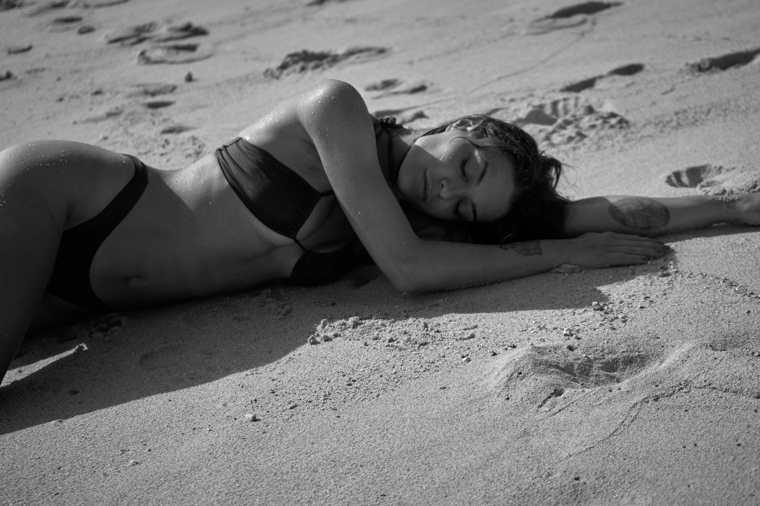 A woman with tattoos and wet hair lying on a sandy beach with her eyes closed, wearing a black bikini.