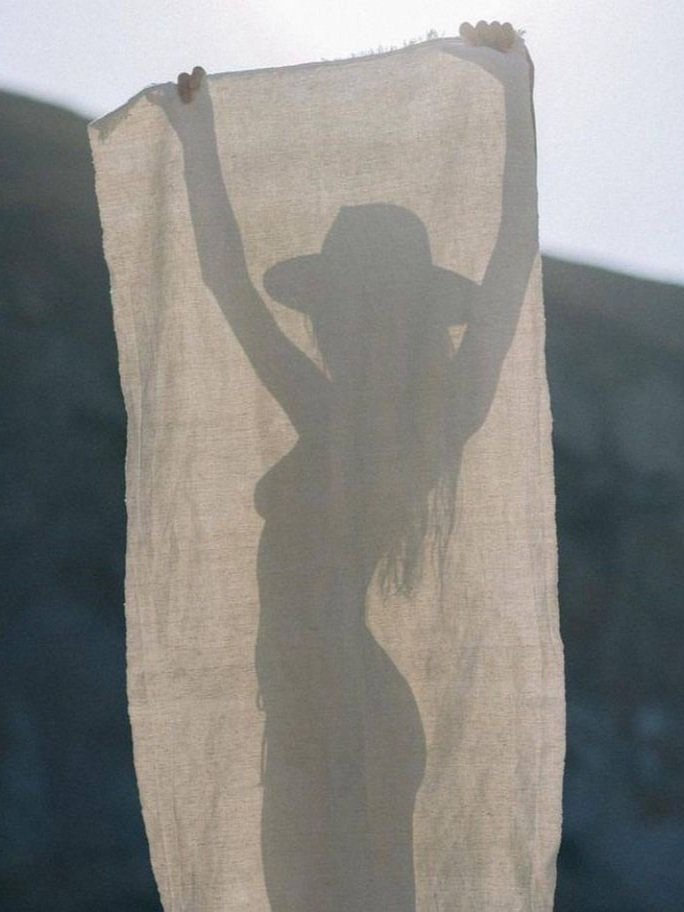 Silhouette of a woman with a hat holding a fabric or paper in front of a light source, creating a shadow outline of her figure.