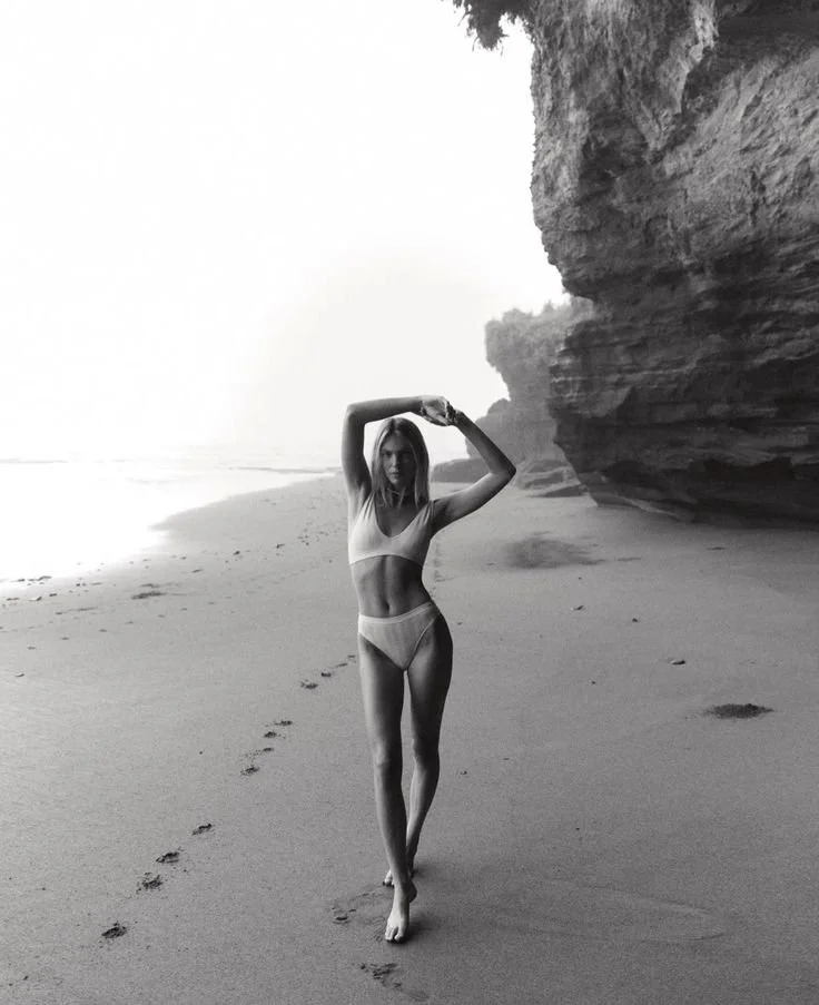 Black and white photograph of a woman in a bikini walking along a beach with footprints trailing behind her, near large rock formations.