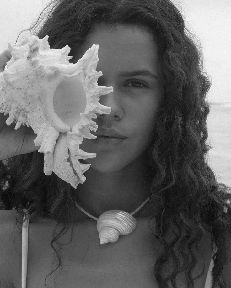 A woman with long curly hair holding a large seashell close to her eye. The photo is in black and white.