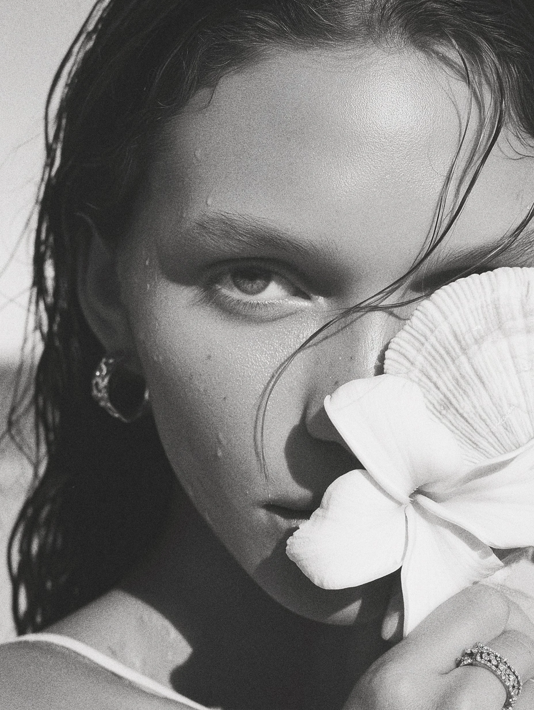 A black and white close-up photo of a woman with wet hair, holding a large flower near her face, partially covering her nose and mouth, with her visible eye gazing at the camera.