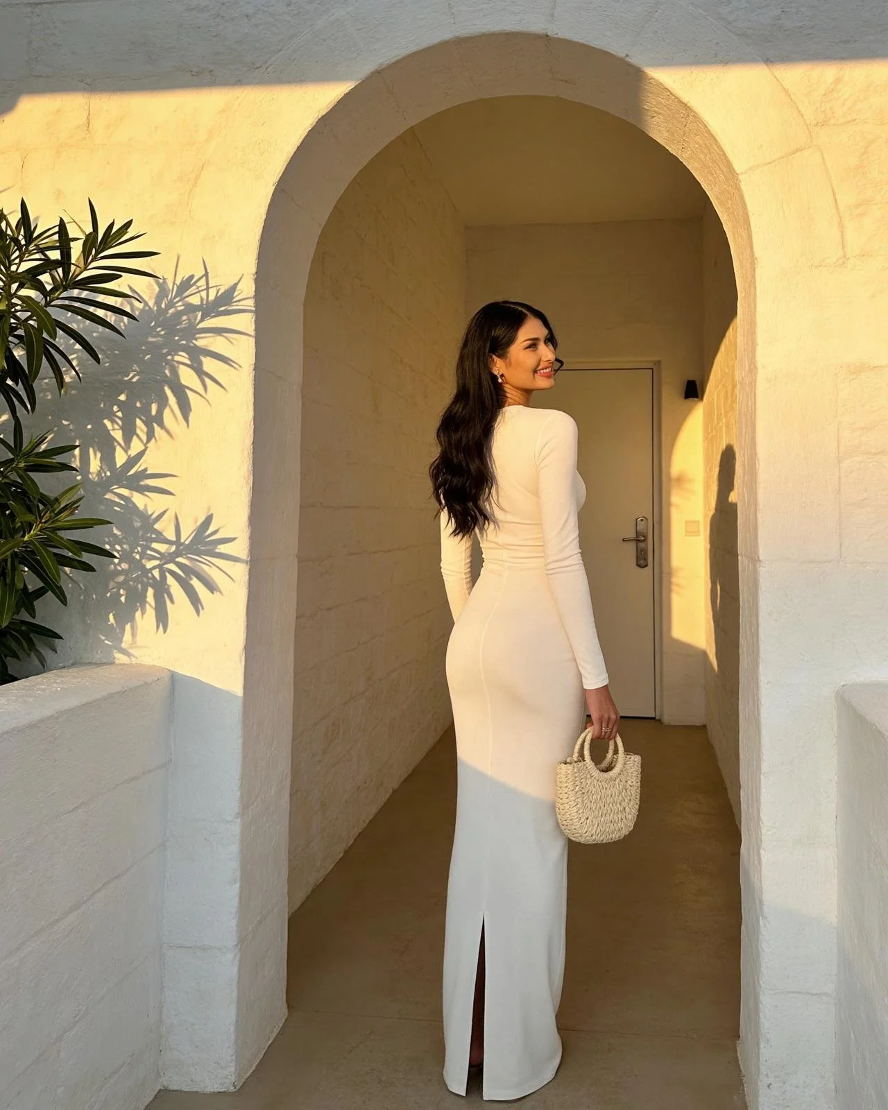 A woman in a long white dress standing in a beige stone archway, smiling, holding a beige woven handbag, with sunlight casting shadows on the wall.