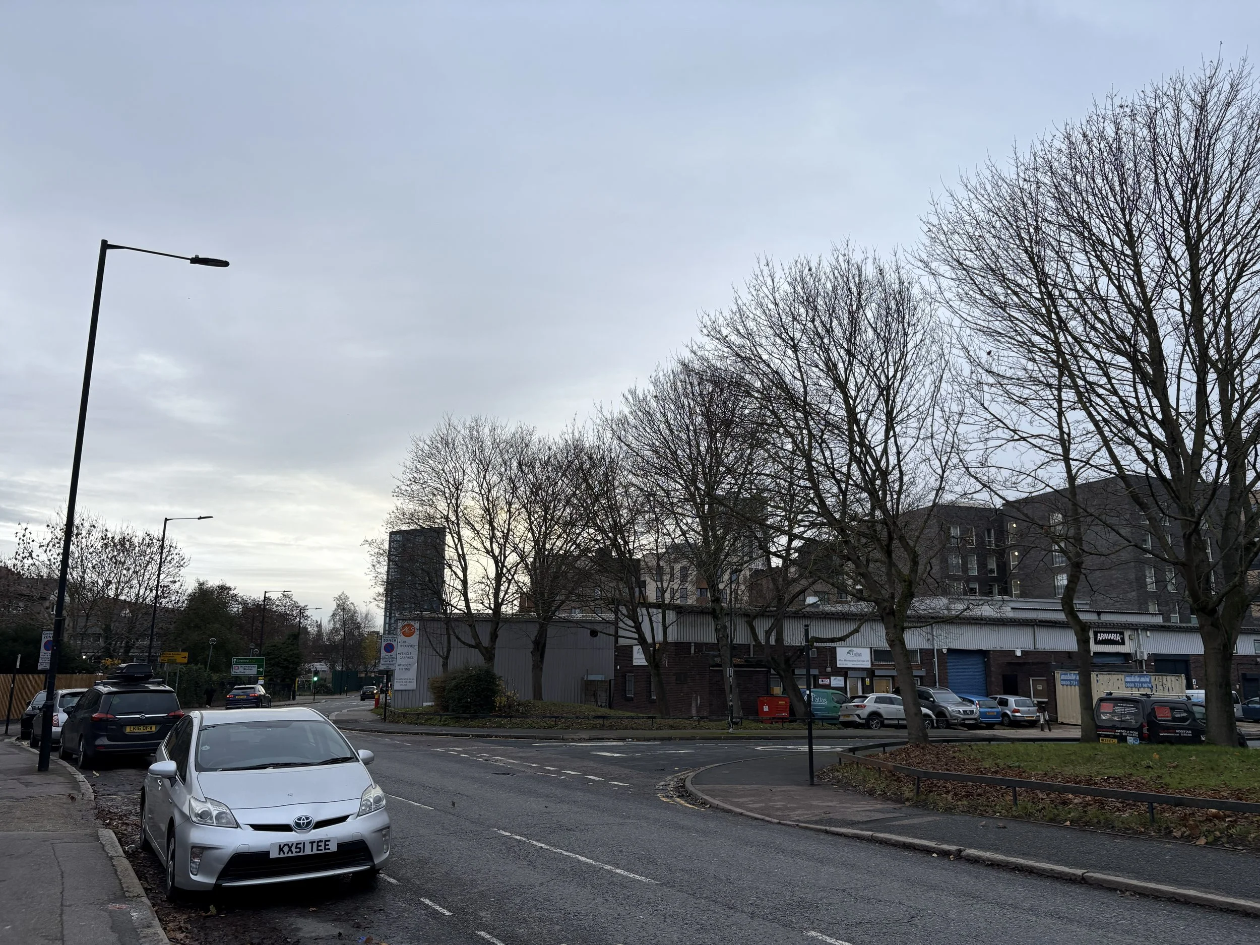 A street scene with parked cars, leafless trees, and a small commercial building in the background under a cloudy sky.