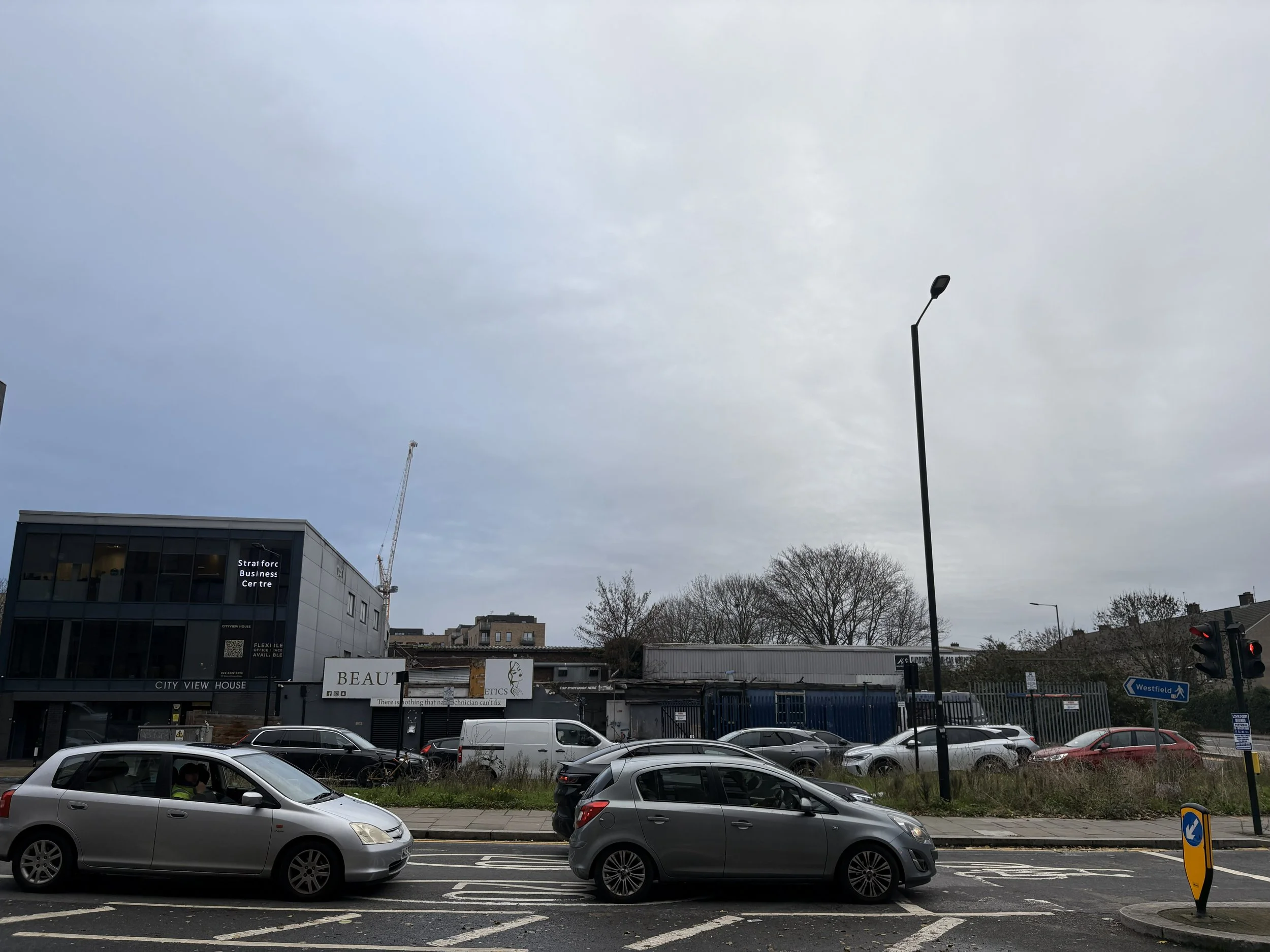 Street scene with cars in traffic, modern buildings, a construction crane, leafless trees, and overcast sky.