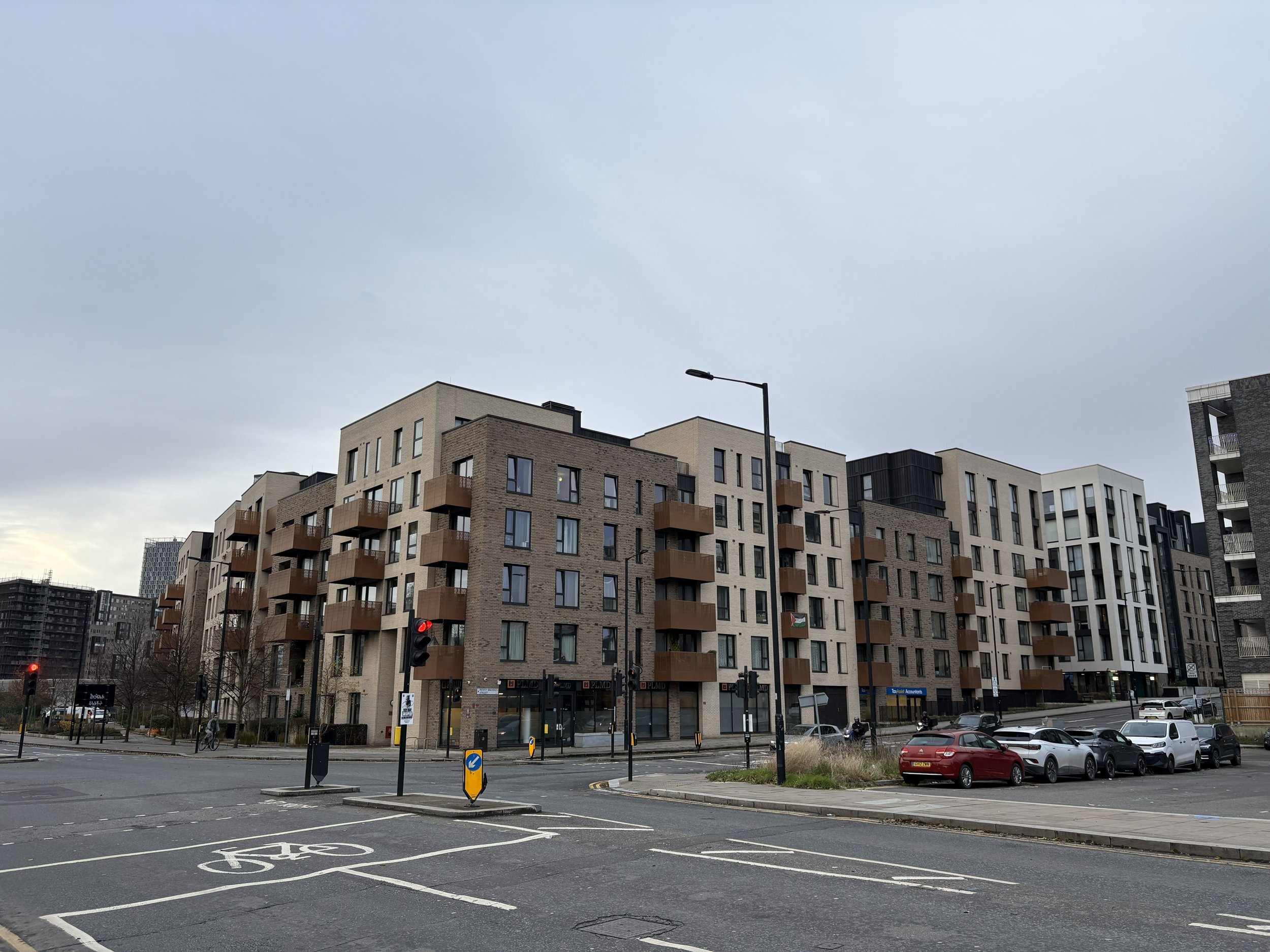 Modern multi-story apartment building with balconies, surrounded by a parking lot, traffic lights, and a cloudy sky.