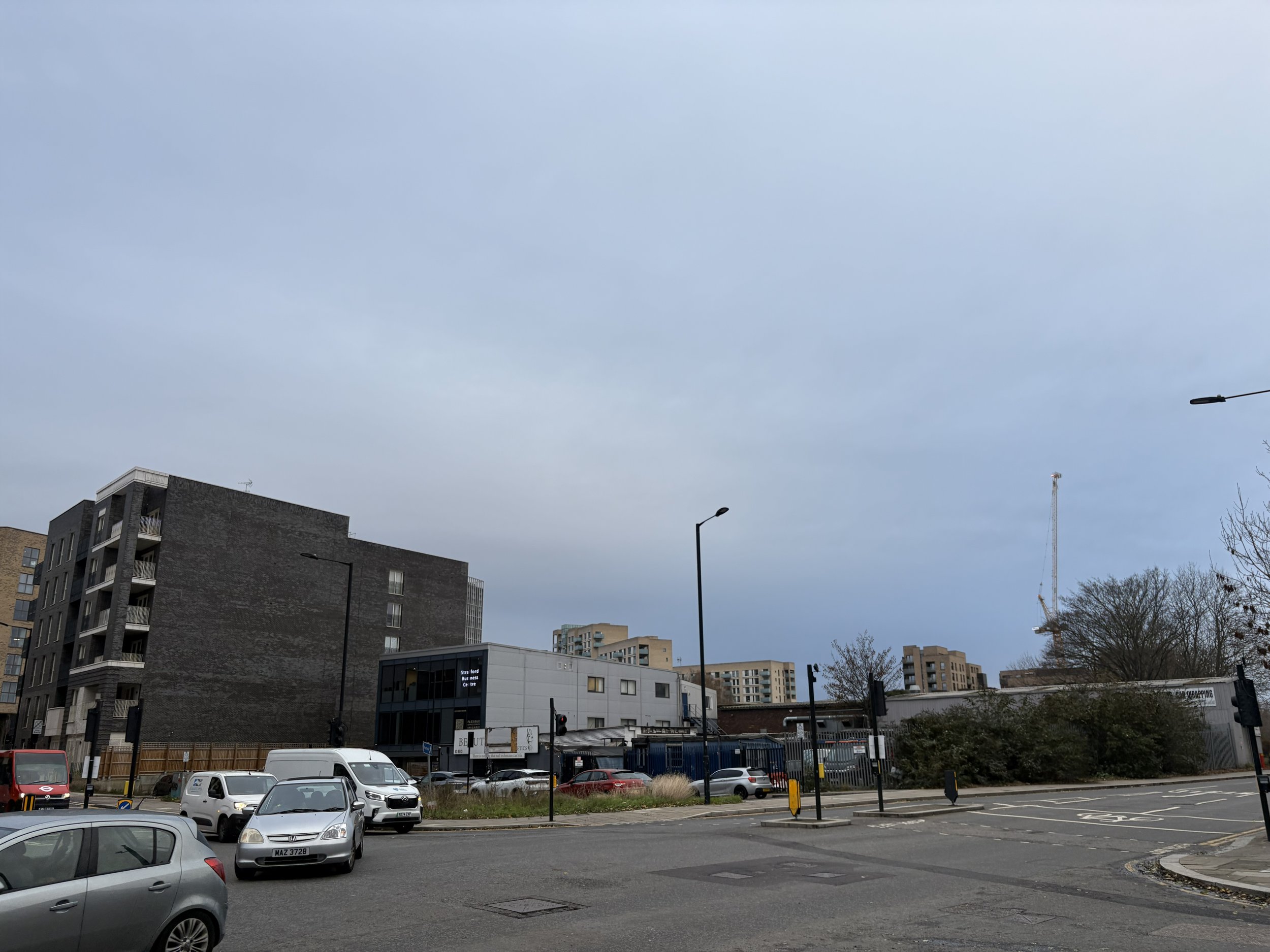 Urban street scene with parked cars, a building with a dark brick facade, utility poles, trees, and a construction crane in the background under a cloudy sky.
