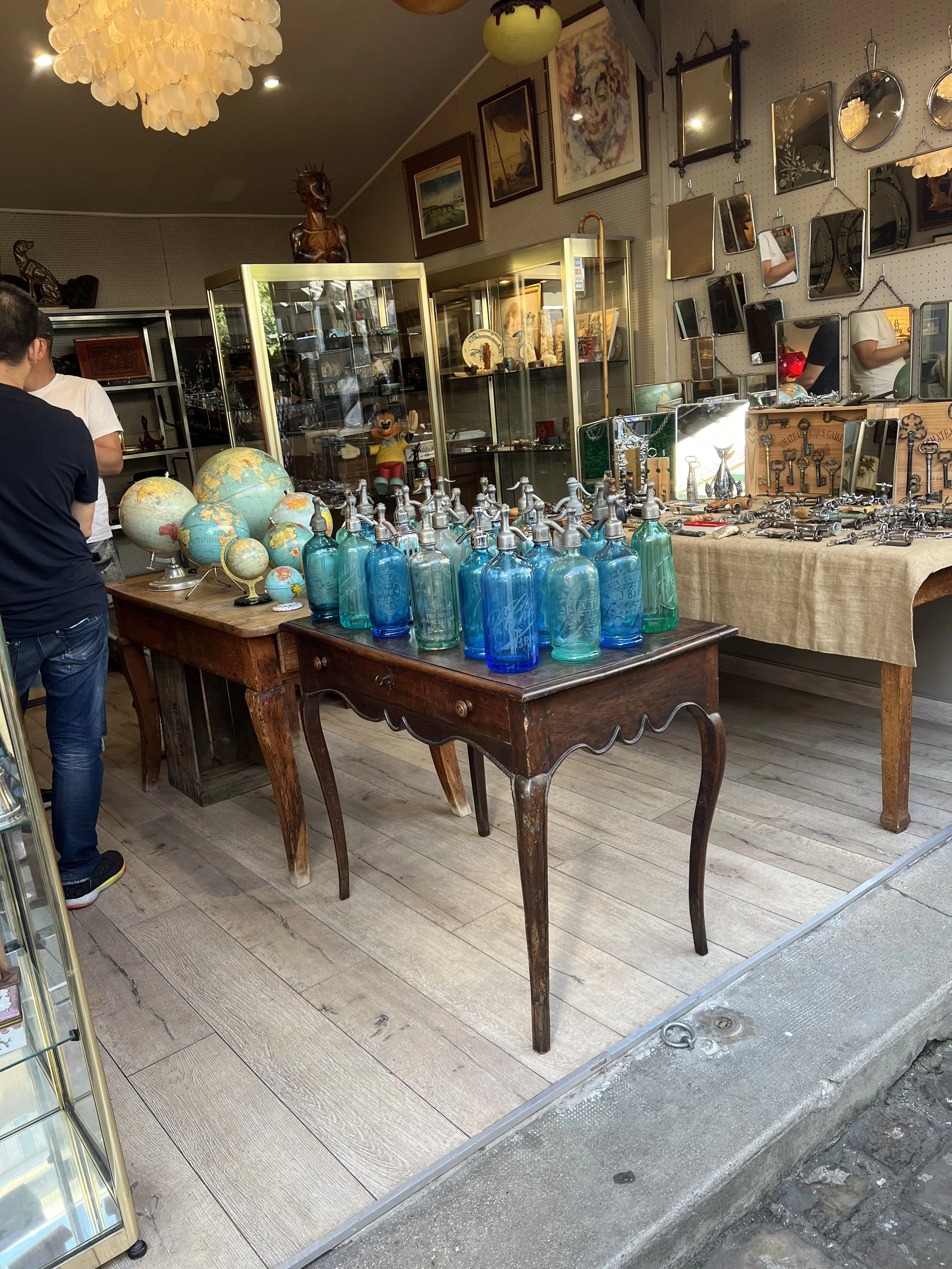 Antique store display with blue glass bottles on a wooden table, globes, and various collectible items, framed artwork, and mirrors on the walls at the famed St. Ouen des Puces market in Paris.