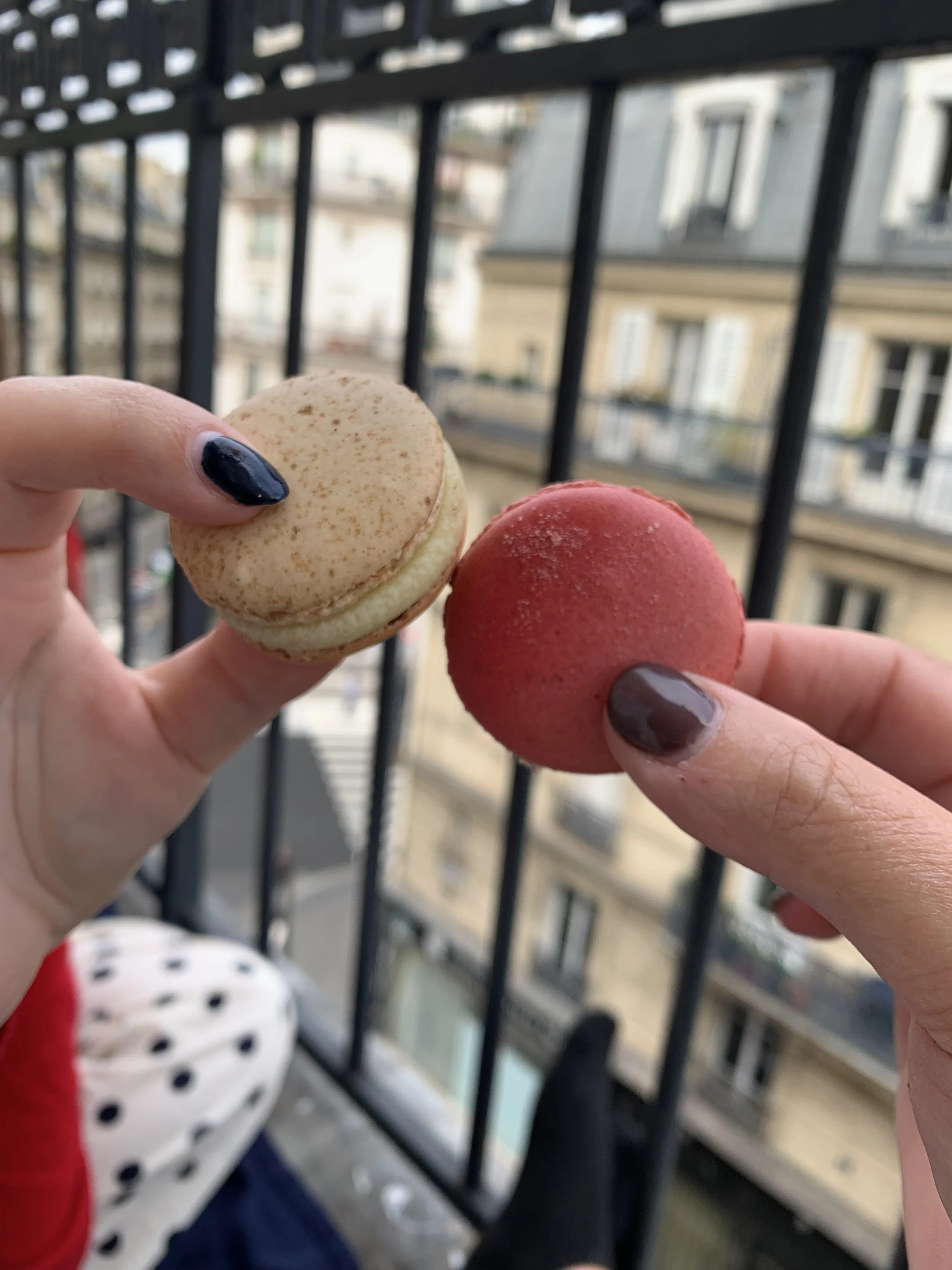 Two macarons—one beige with brown speckles and one pink—held in hands with black nail polish, against an outdoor balcony in St. Germaine Paris
