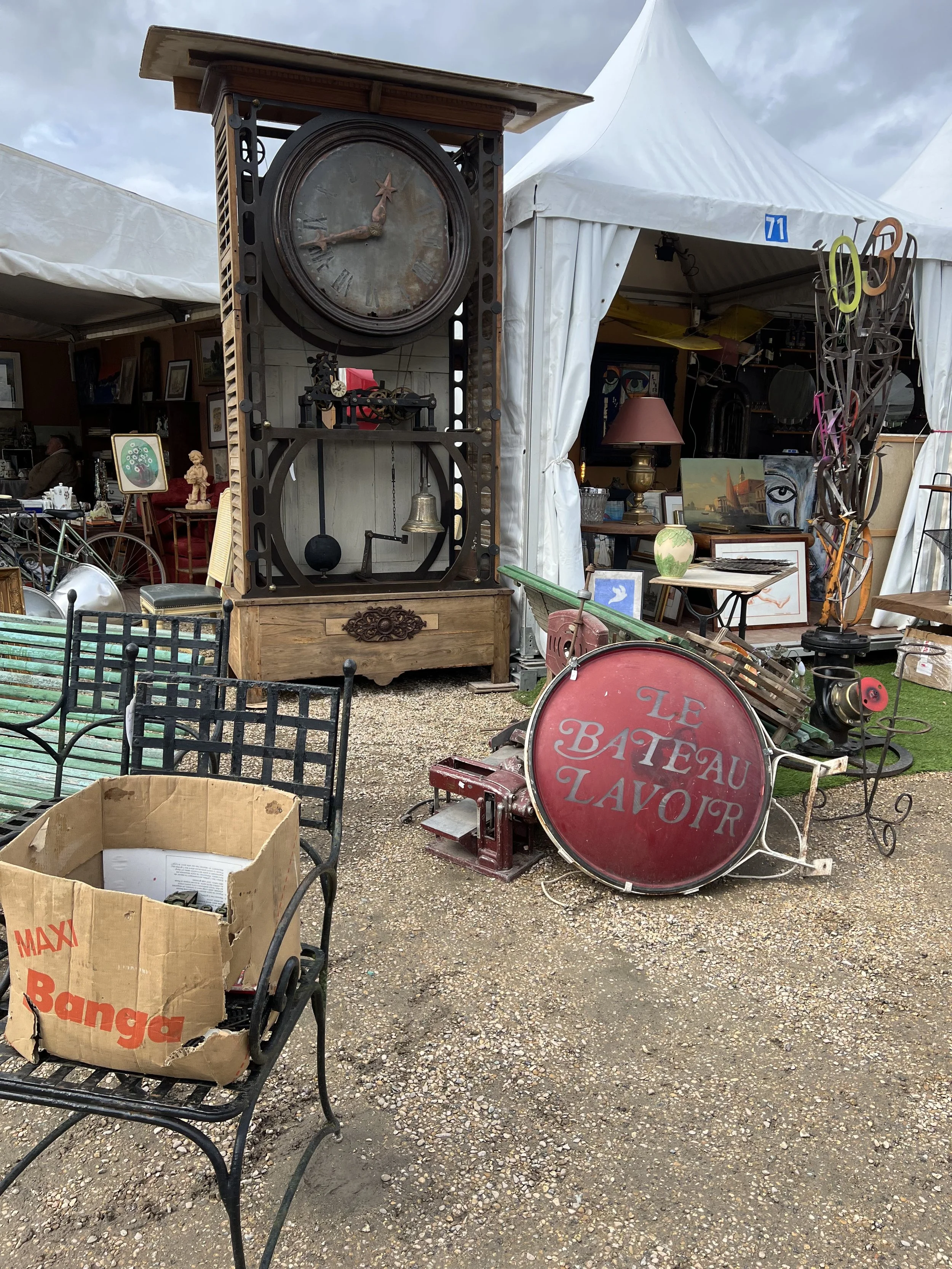 An outdoor antique market in Paris , it features a large vintage clock, a red sign with French text, and various eclectic furniture and decorative items under white tents.