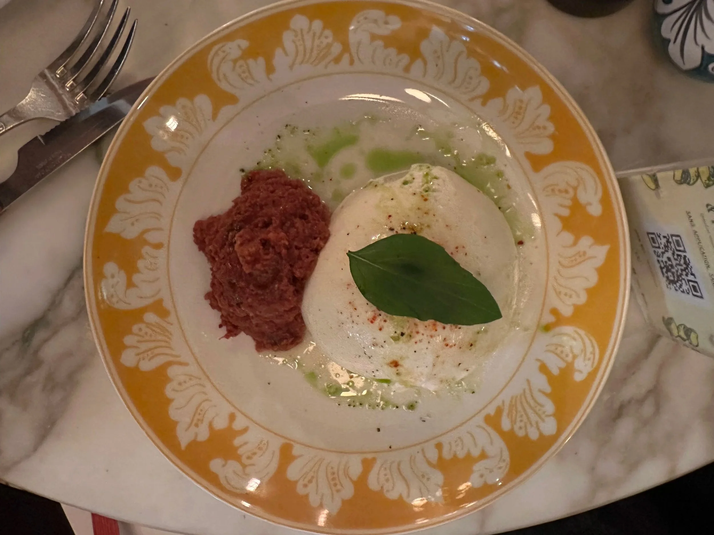 Dish with a scoop of creamy white cheese, a pile of red paste, garnished with a green basil leaf, served on a decorative yellow and white rimmed plate.