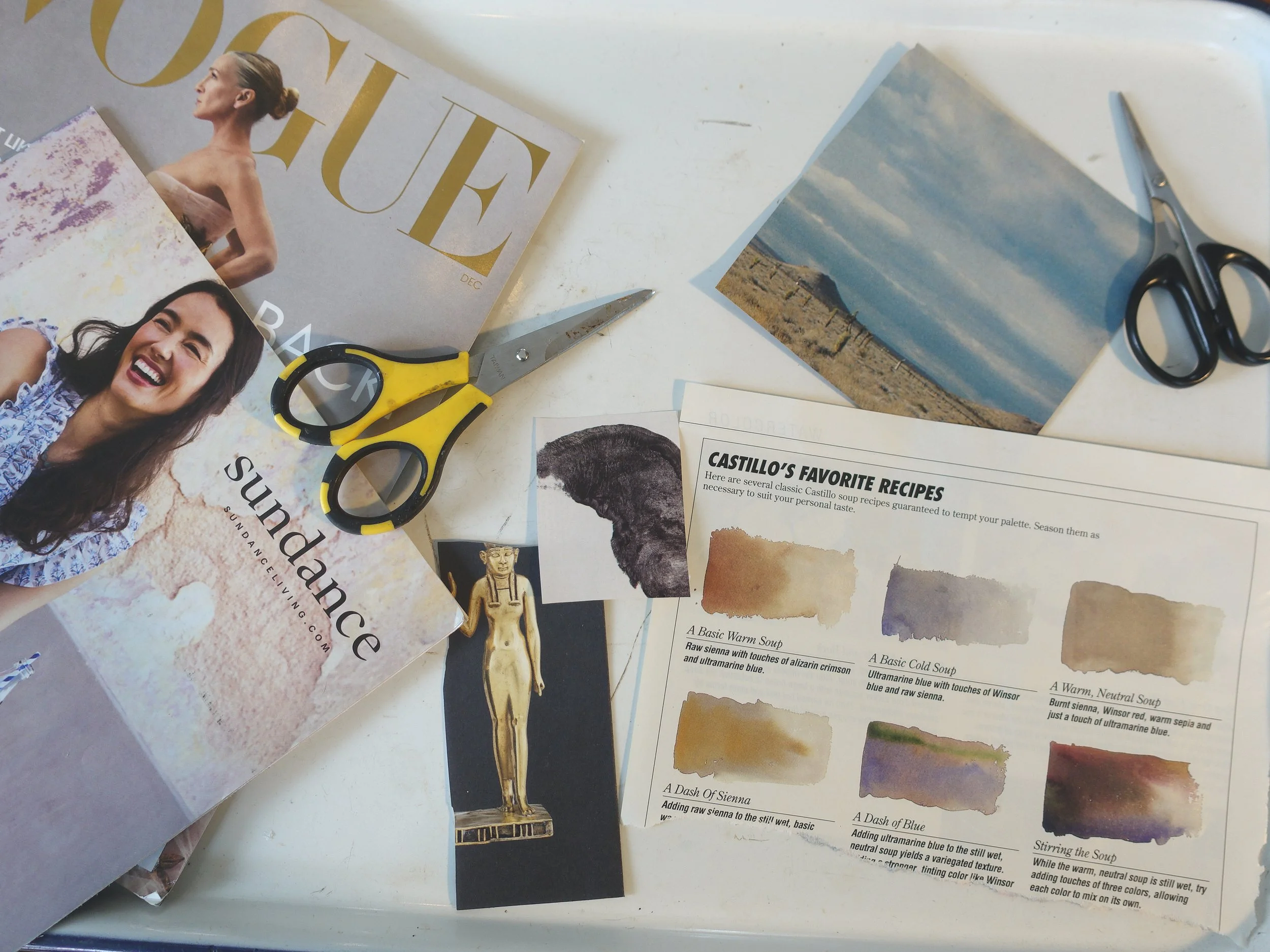 A tabletop with scissors, a magazine titled "Sunday," a photograph of a landscape, a printed page of soup recipes, a small cutout of a woman in a golden dress, and a pair of black scissors.