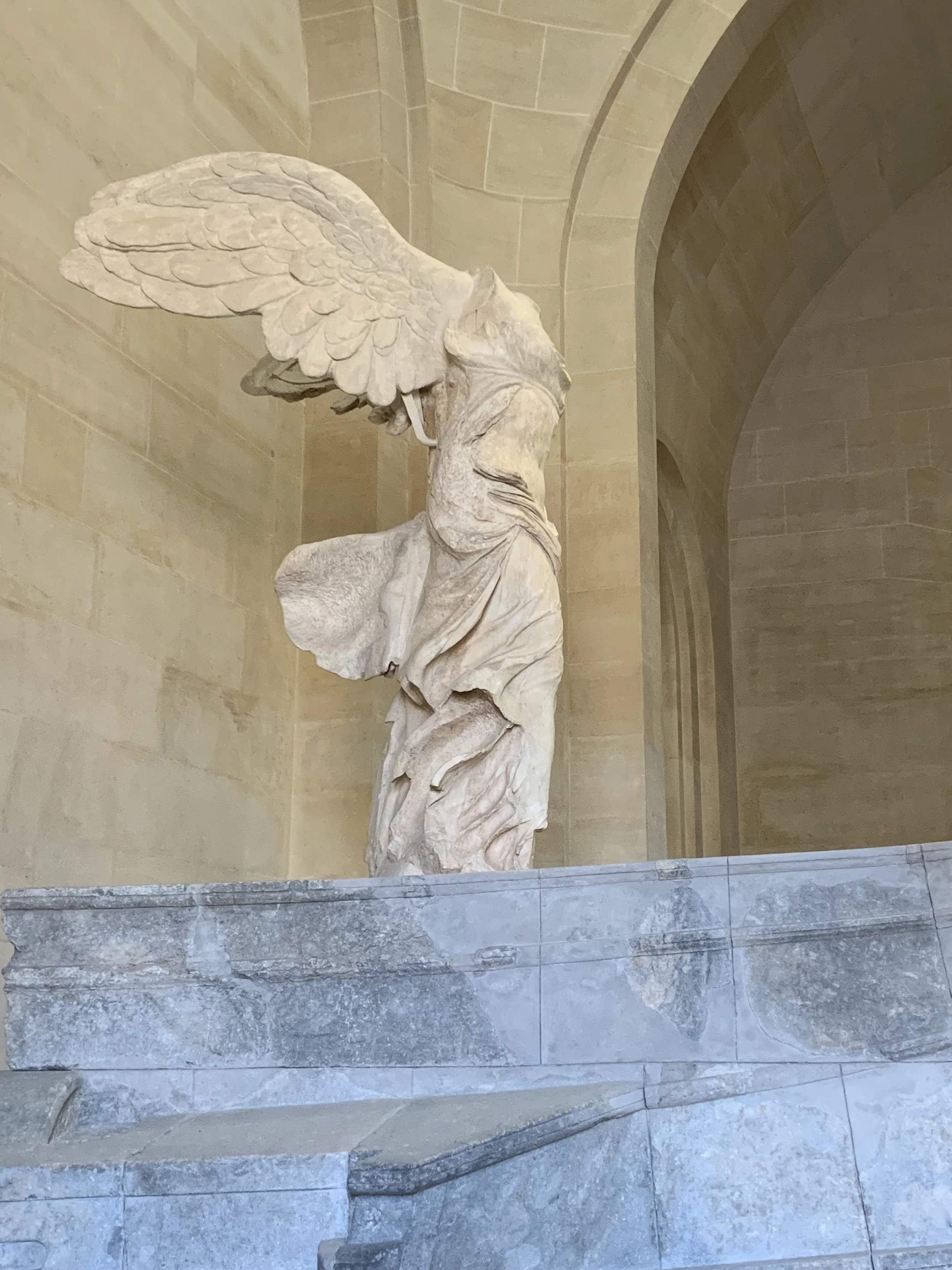 Marble sculpture of a winged figure standing on a stone base in an indoor setting with arched walls.