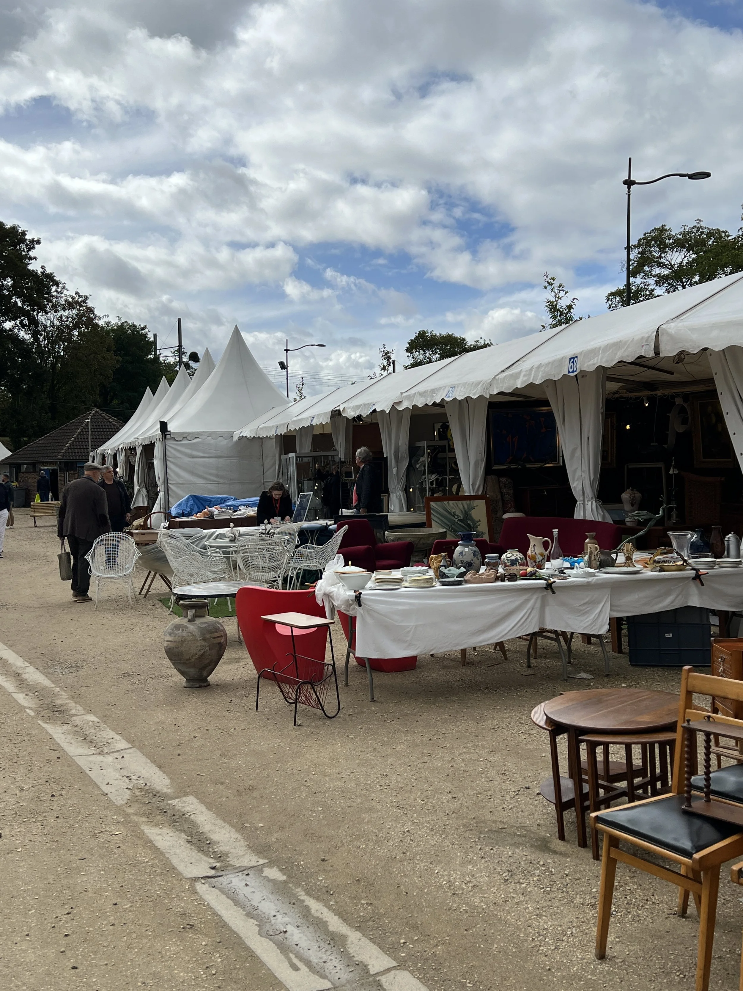 Outdoor flea market In Paris with white tents, tables displaying various items, and people browsing; chairs and decorative objects are visible.