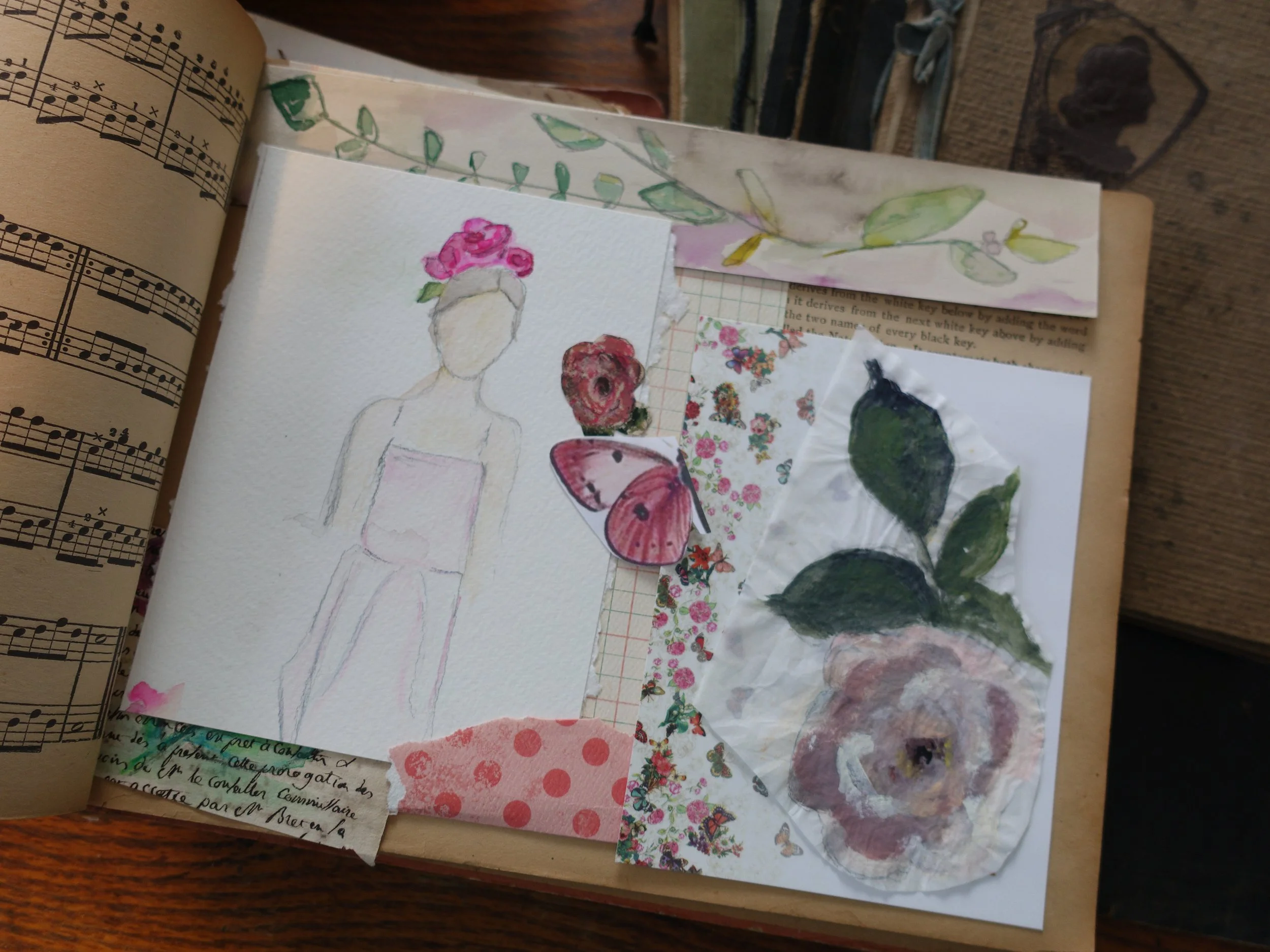 Open sketchbook with a watercolor sketch of a faceless woman wearing a pink dress and pink flowers in her hair. Surrounding the sketch are various floral and butterfly stickers, torn paper pieces with floral designs, and a leaf painting on a crumpled paper, all in a mixed-media art style. The background includes a piece of sheet music and some handwritten notes.