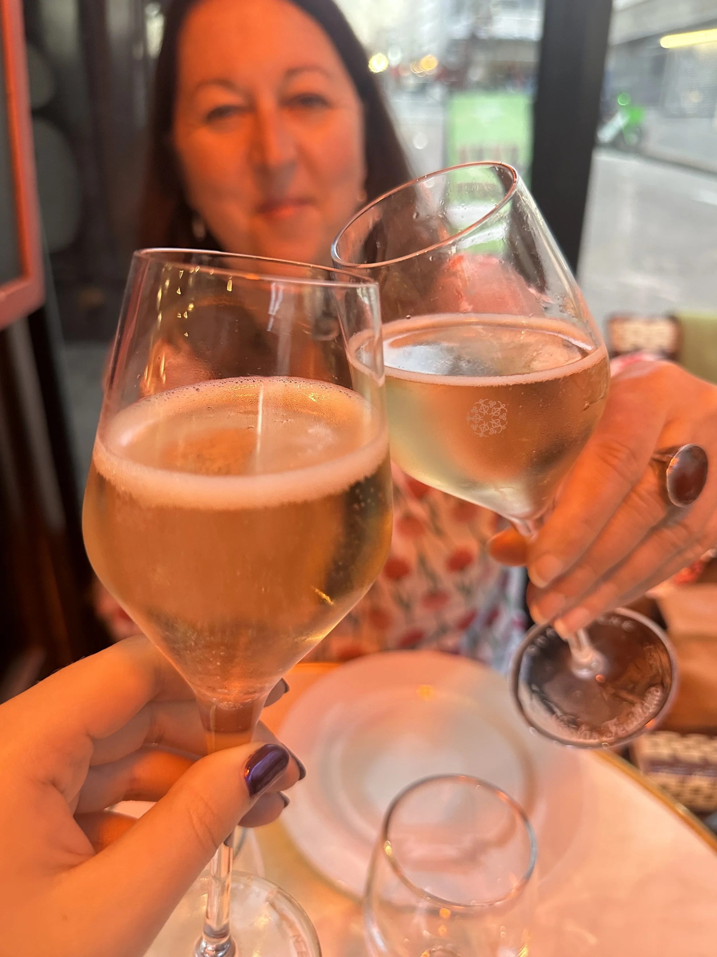 Two women clinking glasses of white wine or champagne in a restaurant, with a woman smiling in the background.