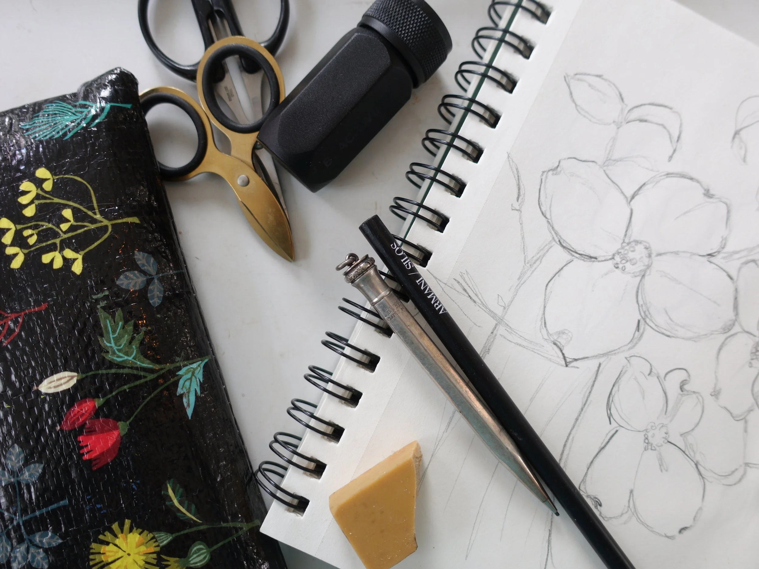 Art supplies on a white surface including a sketchbook with a floral drawing, pencils, a black glue stick, a block of beeswax, scissors, a black spray bottle, and a black bag with colorful floral designs.