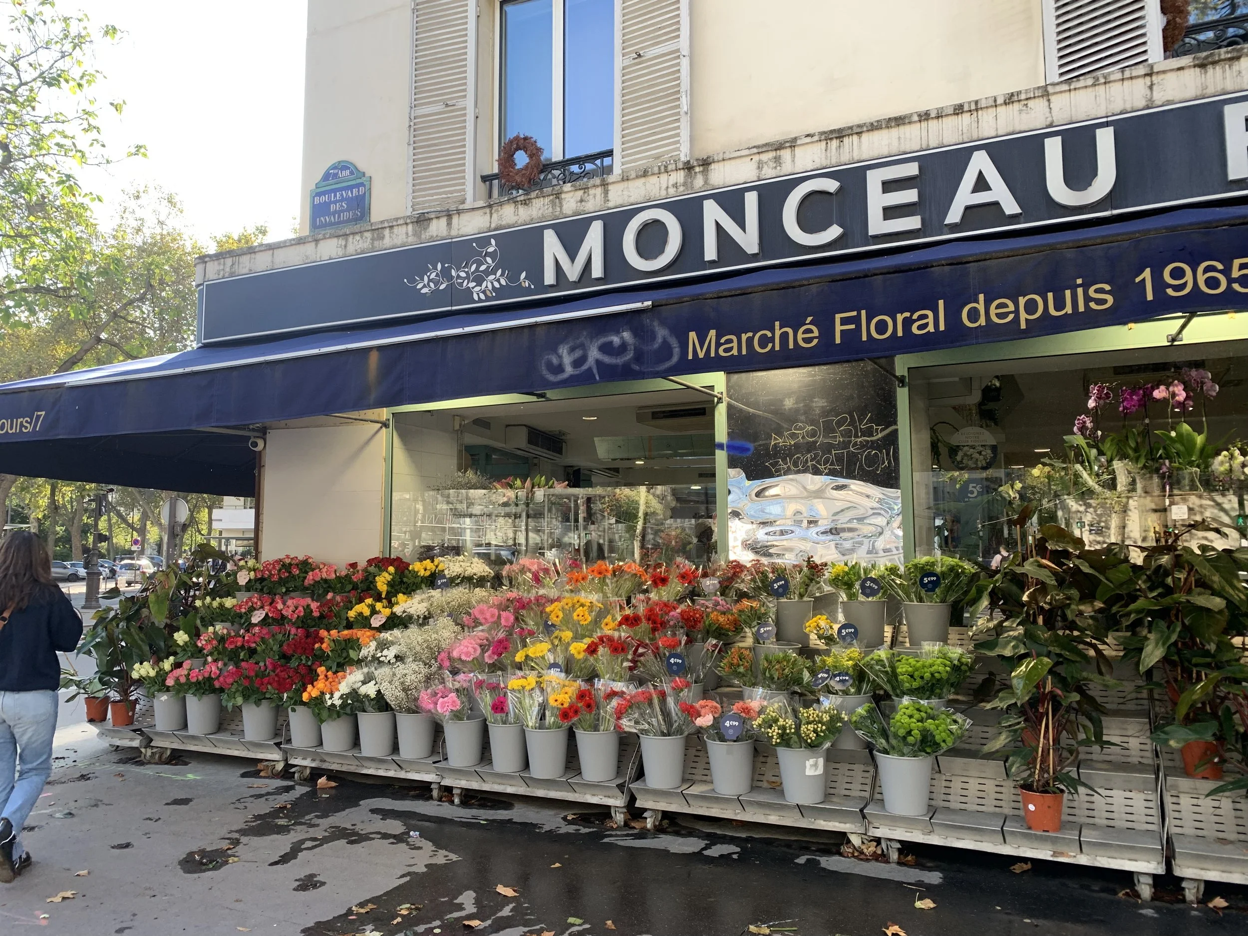 Flower shop with colorful potted flowers on display outside, with a blue awning reading 'Monceau' and 'Marché Floral depuis 1965', located on a street in Paris.