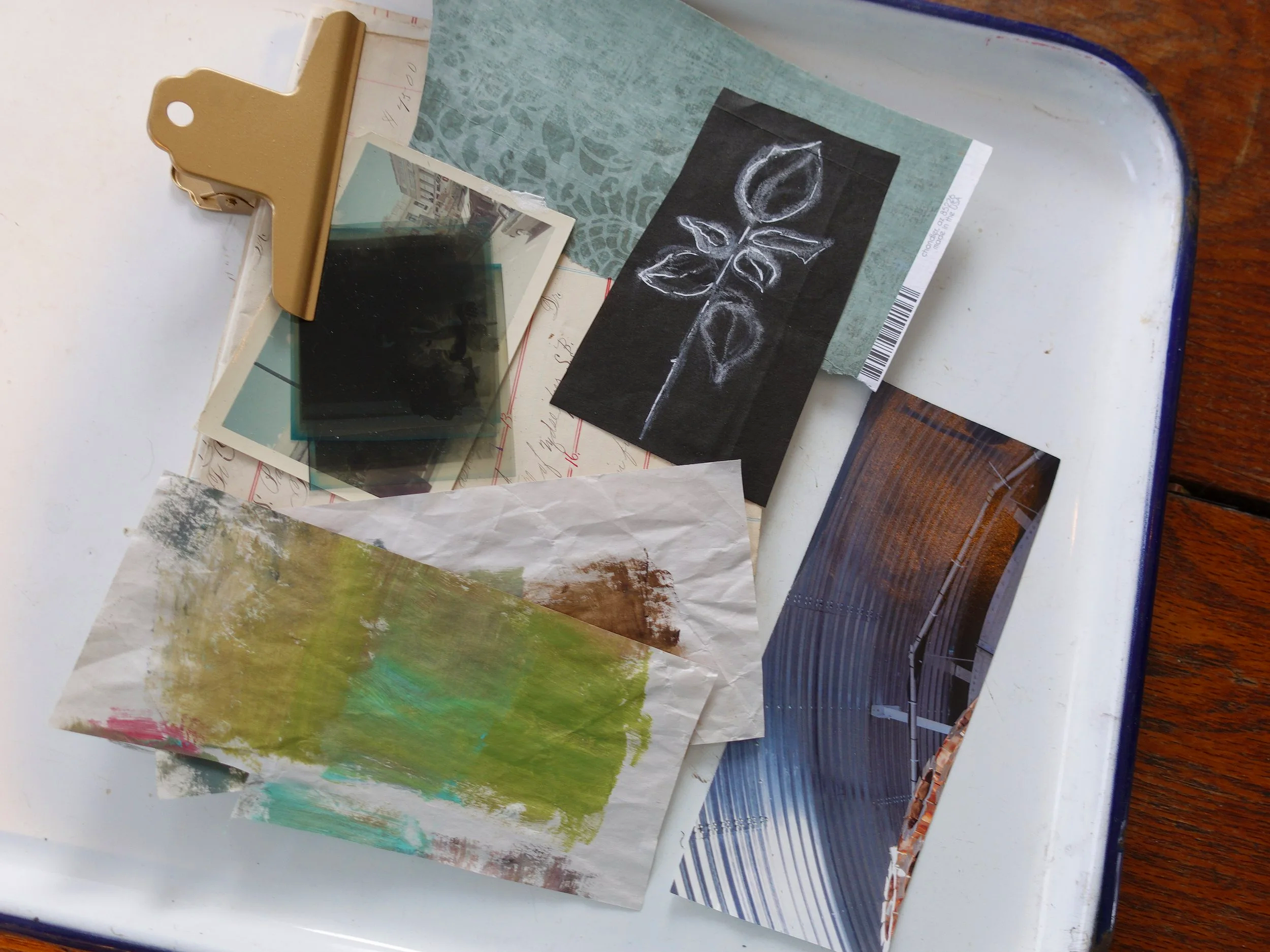 A collection of small photographs, artwork, and paper samples on a white tray, with a gold clip holding some items, on a wooden surface.