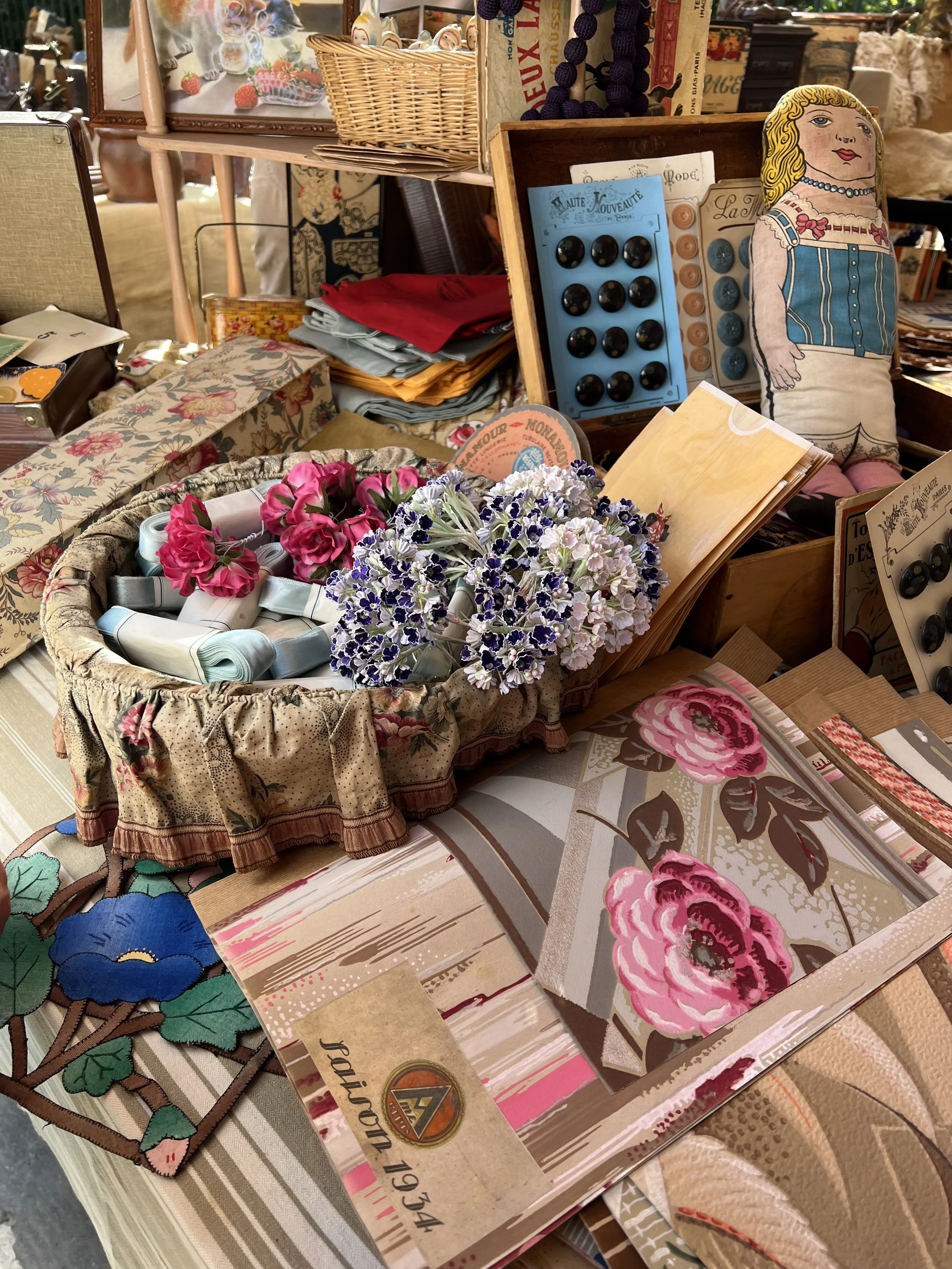 A cluttered display of vintage textiles, decorative items, and craft supplies. Items include a floral fabric box with artificial flowers, patterned fabrics, a doll-shaped pillow, buttons, a basket of rolled ribbons, printed fabric swatches, and small