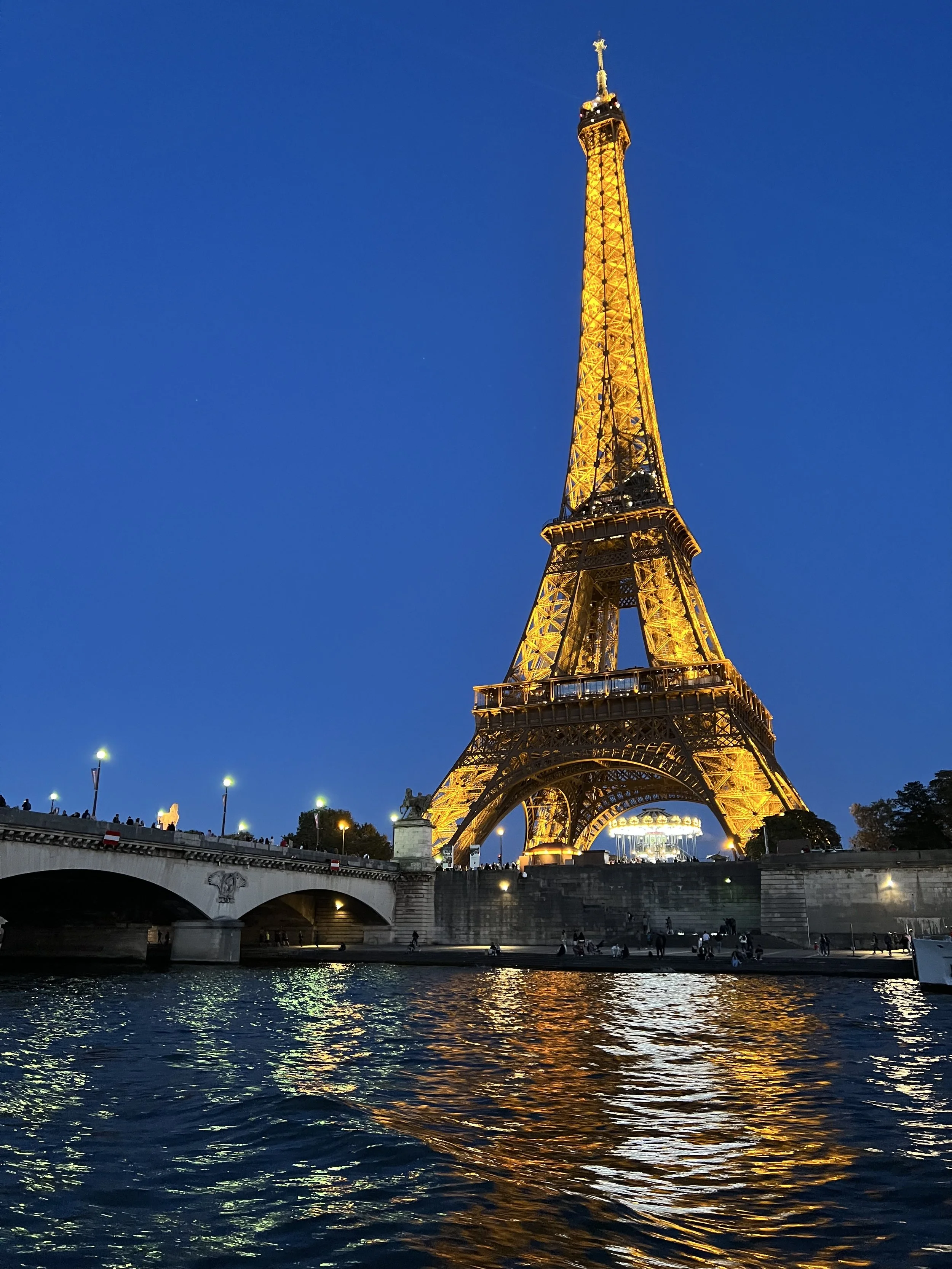 The Eiffel Tower illuminated at night in Paris, viewed from across the river with reflections on the water.