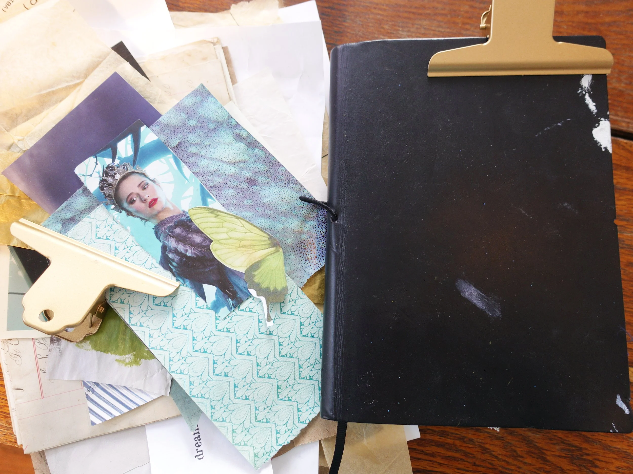 A black notebook with a gold clip on a wooden surface, surrounded by various arts and crafts papers, a cutout image of a woman with a crown, butterfly wings, and decorative paper.