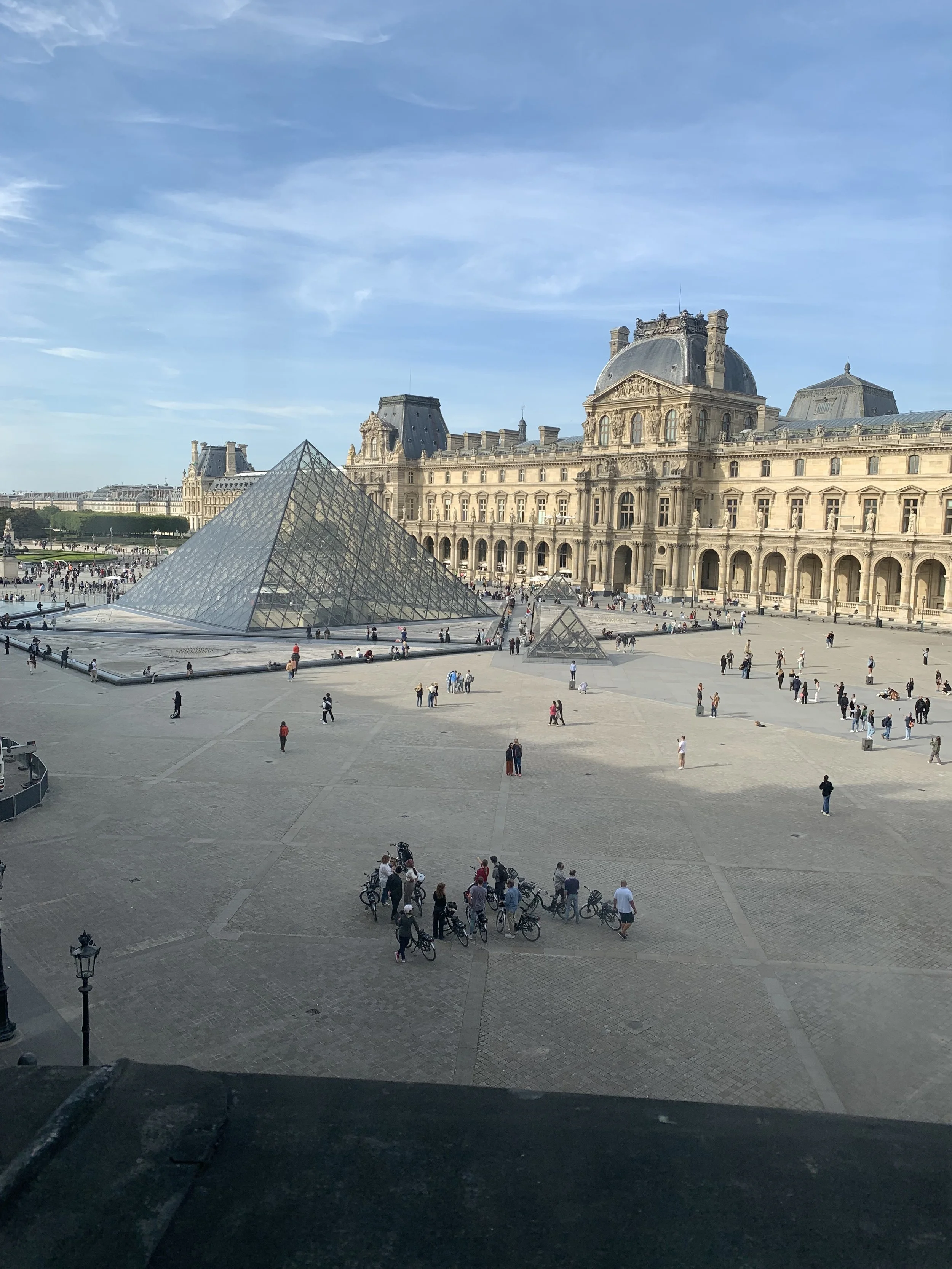 View of the Louvre Museum in Paris, France, including glass pyramid entrances and people walking and cycling in the courtyard.