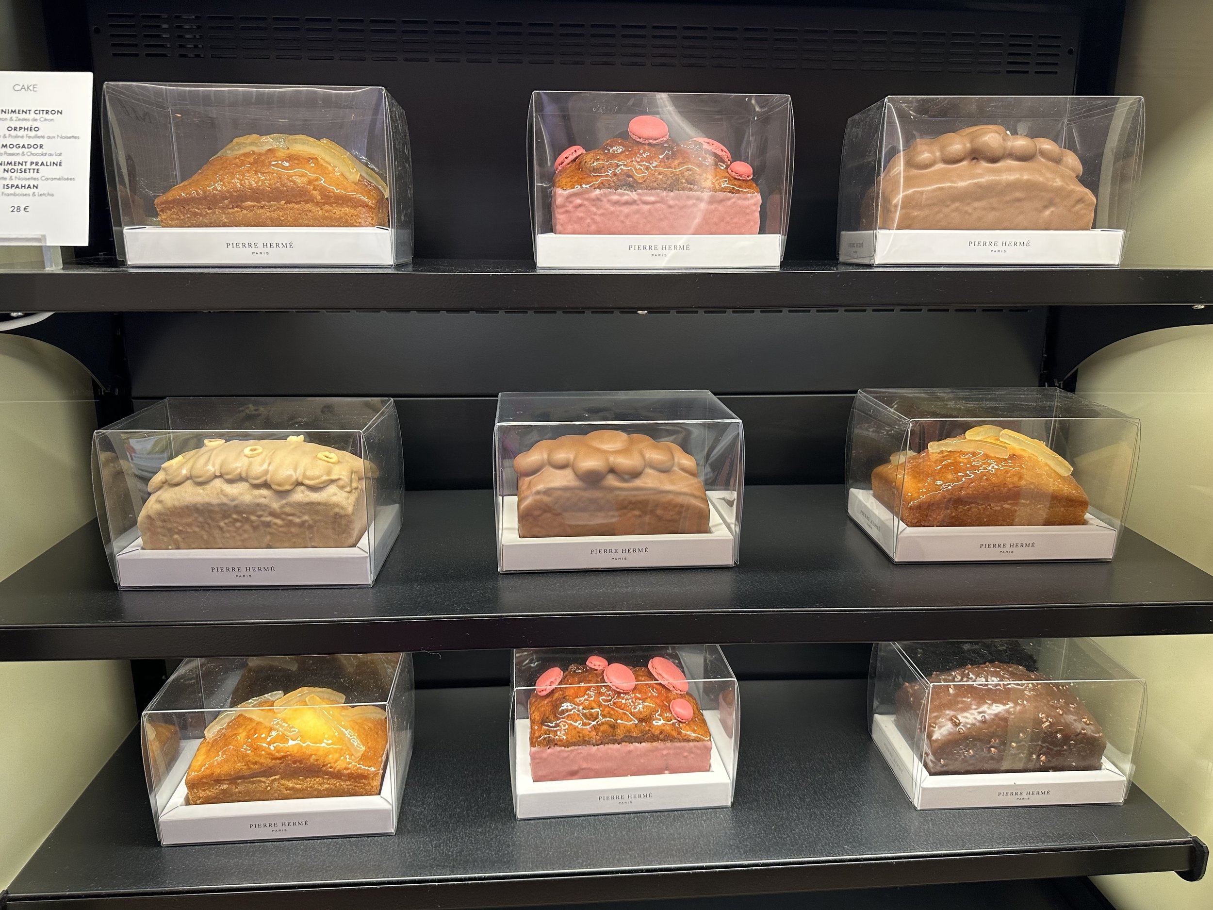 Display of assorted pastries in clear boxes on black shelves at Pierre Hermé bakery.