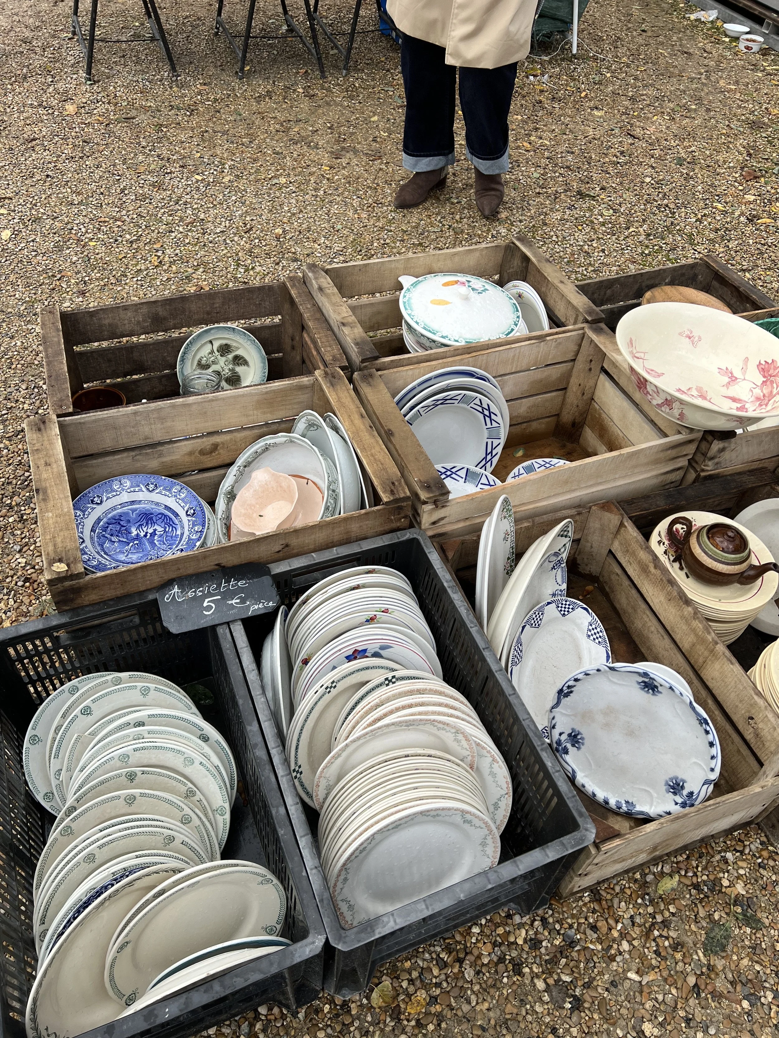 Assorted vintage dishes and plates displayed in wooden crates and black baskets at an outdoor market. A person in a beige coat and brown boots stands behind the display.