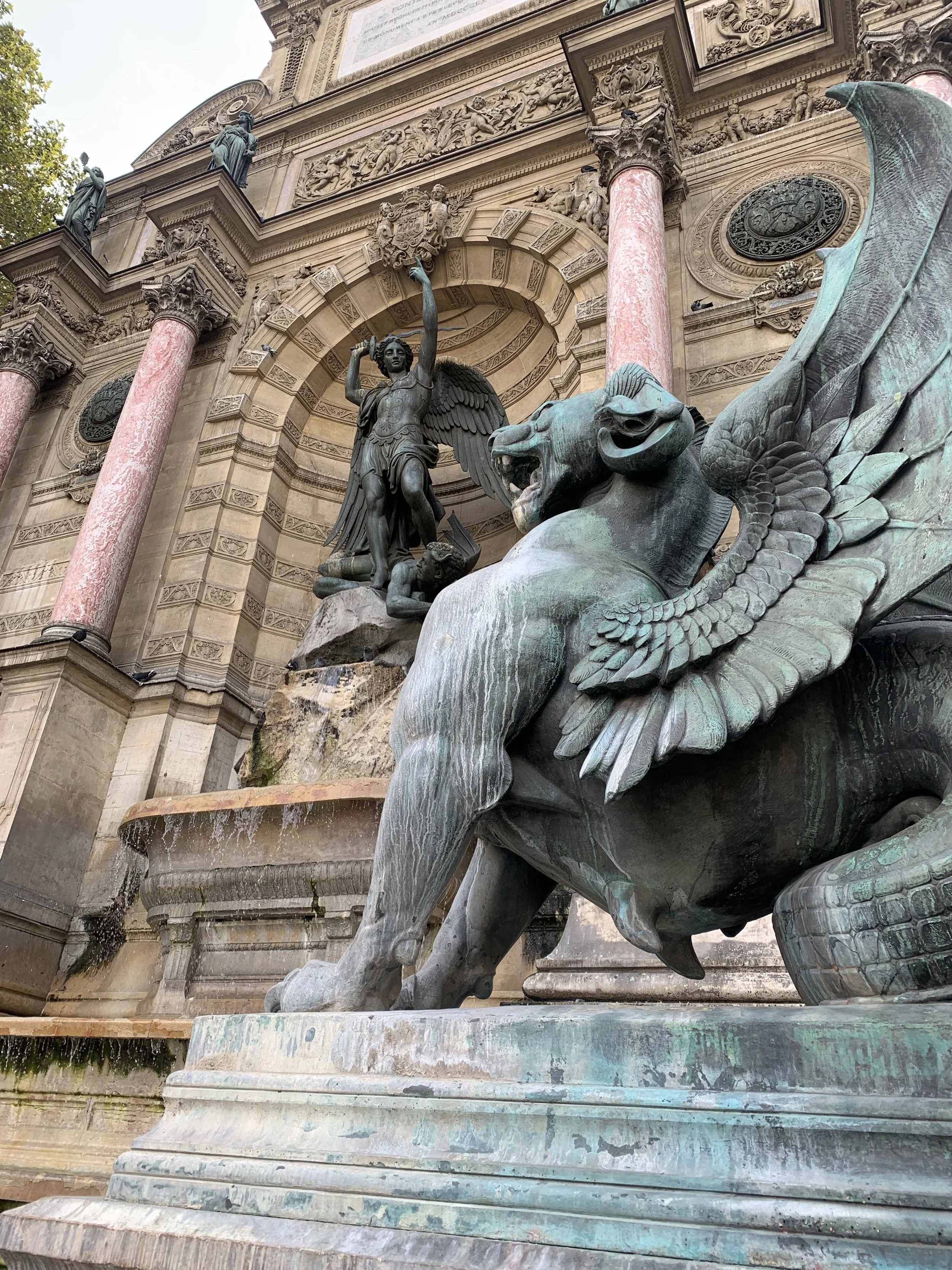 A historical fountain featuring sculptures of a winged figure holding a sword and a shield, and a large winged lion with a commanding stance, set against an ornate building with columns and detailed reliefs.
