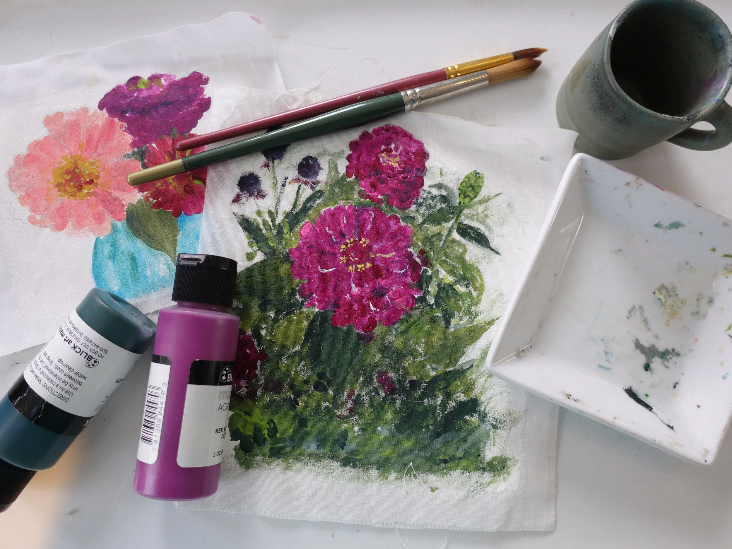 Painted floral artwork on fabric with bottles of purple and teal acrylic paint, paintbrushes, a mug, and a white palette with paint smears.