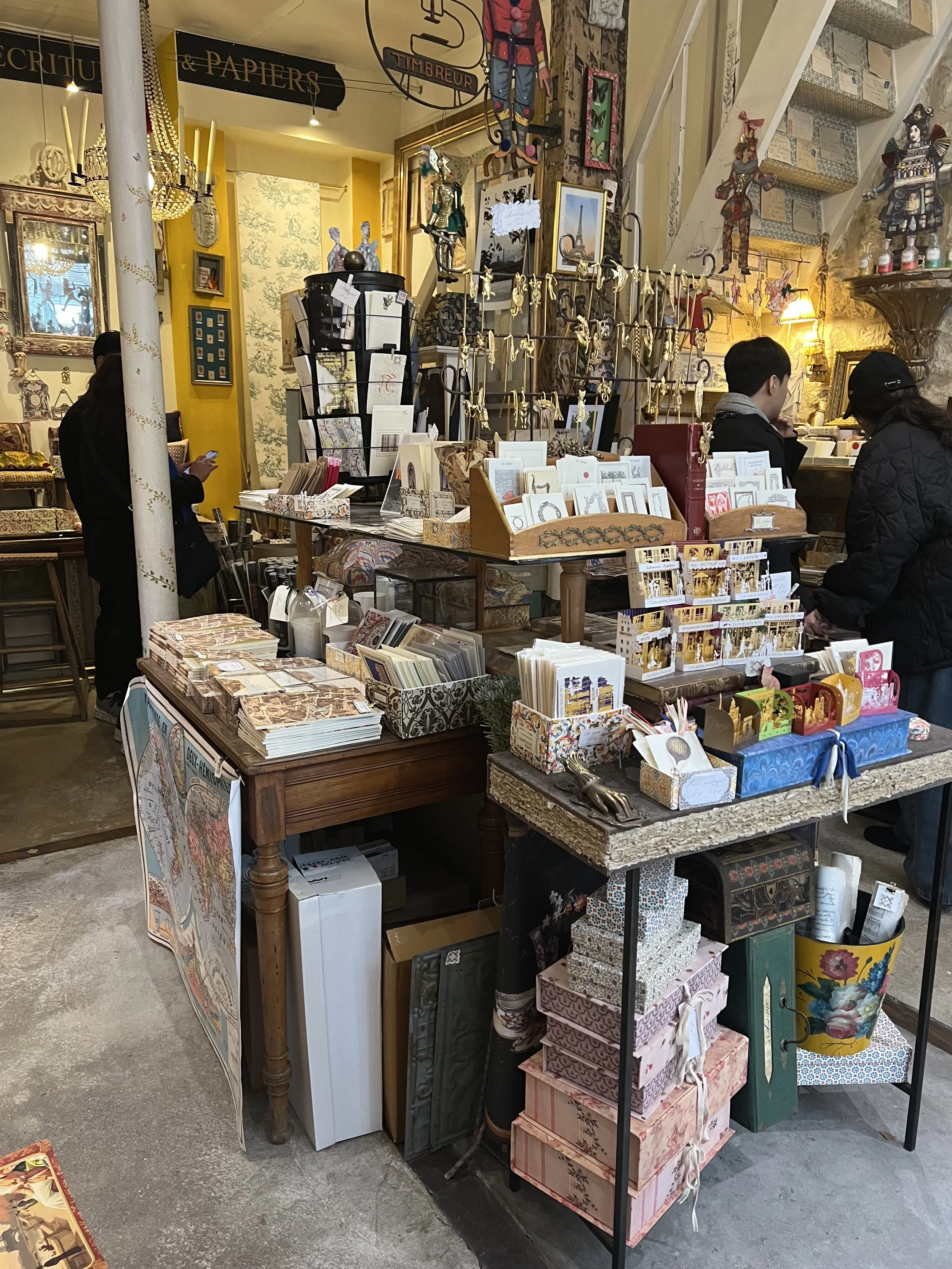 A store with shelves and tables displaying stationery, greeting cards, and decorative items, with customers browsing inside at the Marche aux Puces Paris