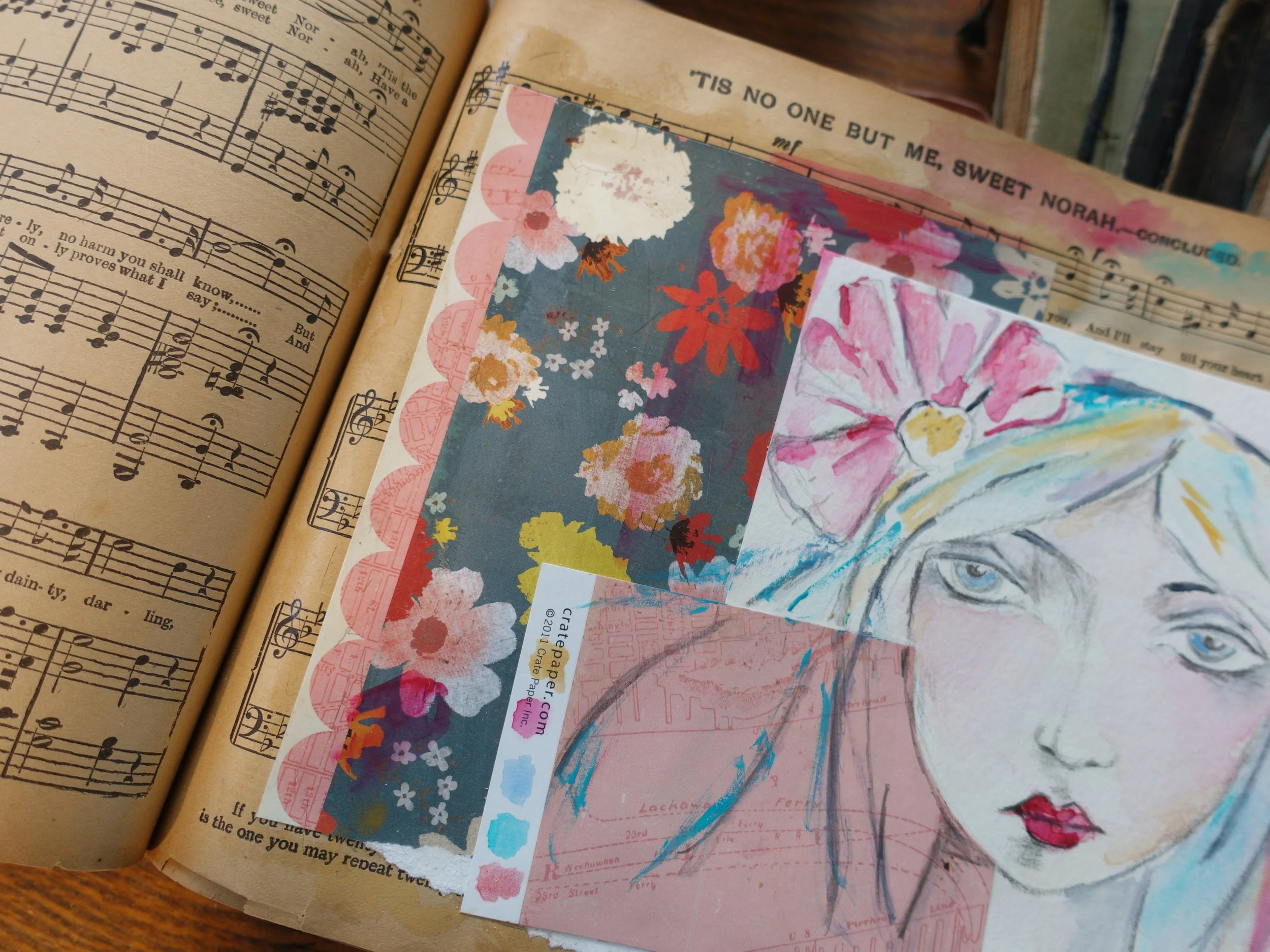 Open sheet music book alongside colorful artistic sketches and cutouts on paper, including a portrait of a girl with light hair and blue eyes, and floral patterns.