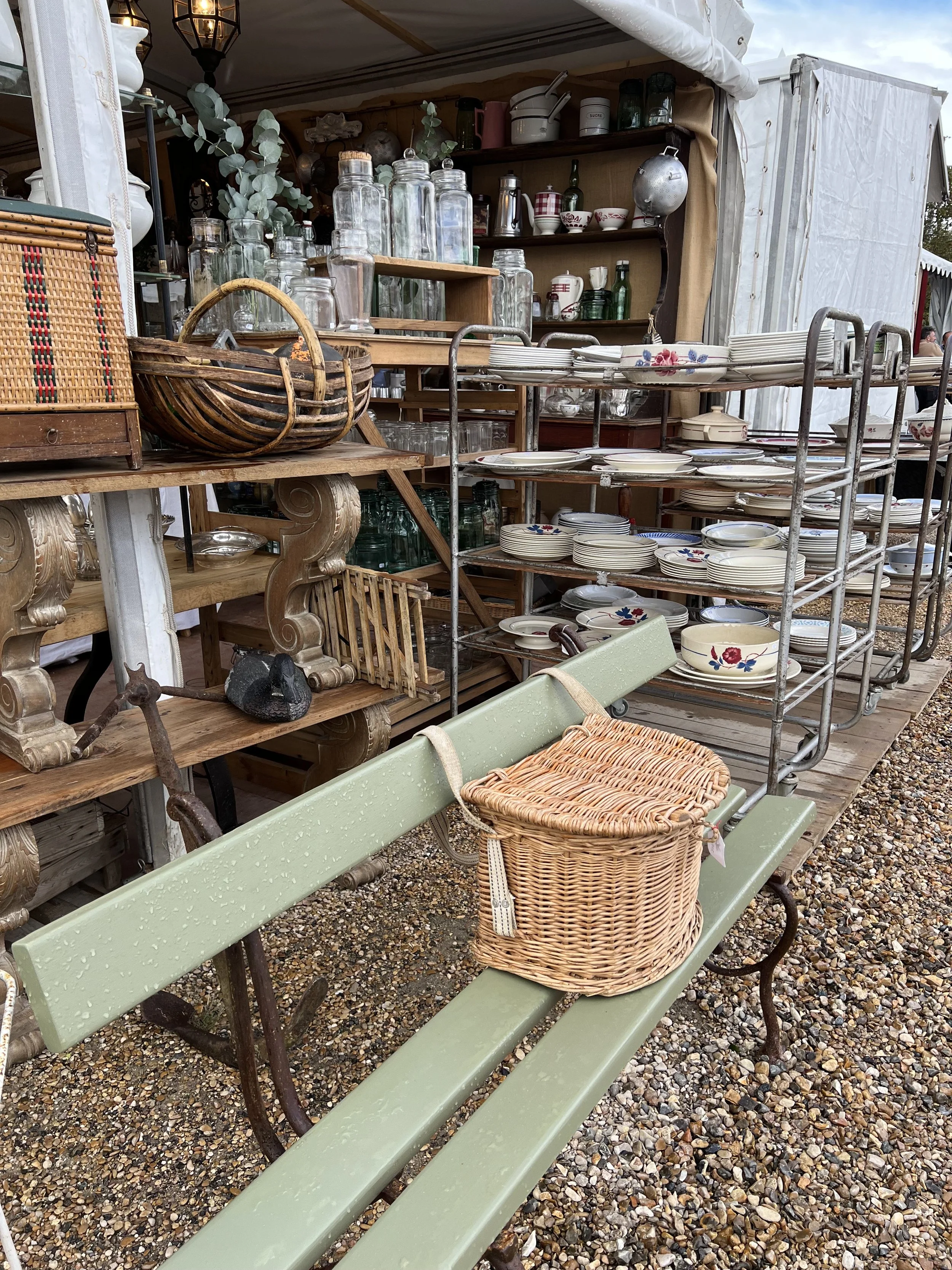A green park bench with raindrops on it, placed on gravel, with a wicker basket on top, and at a Paris vintage outdoor market with shelves of glass jars, plates, bowls, and various antique items in the background.
