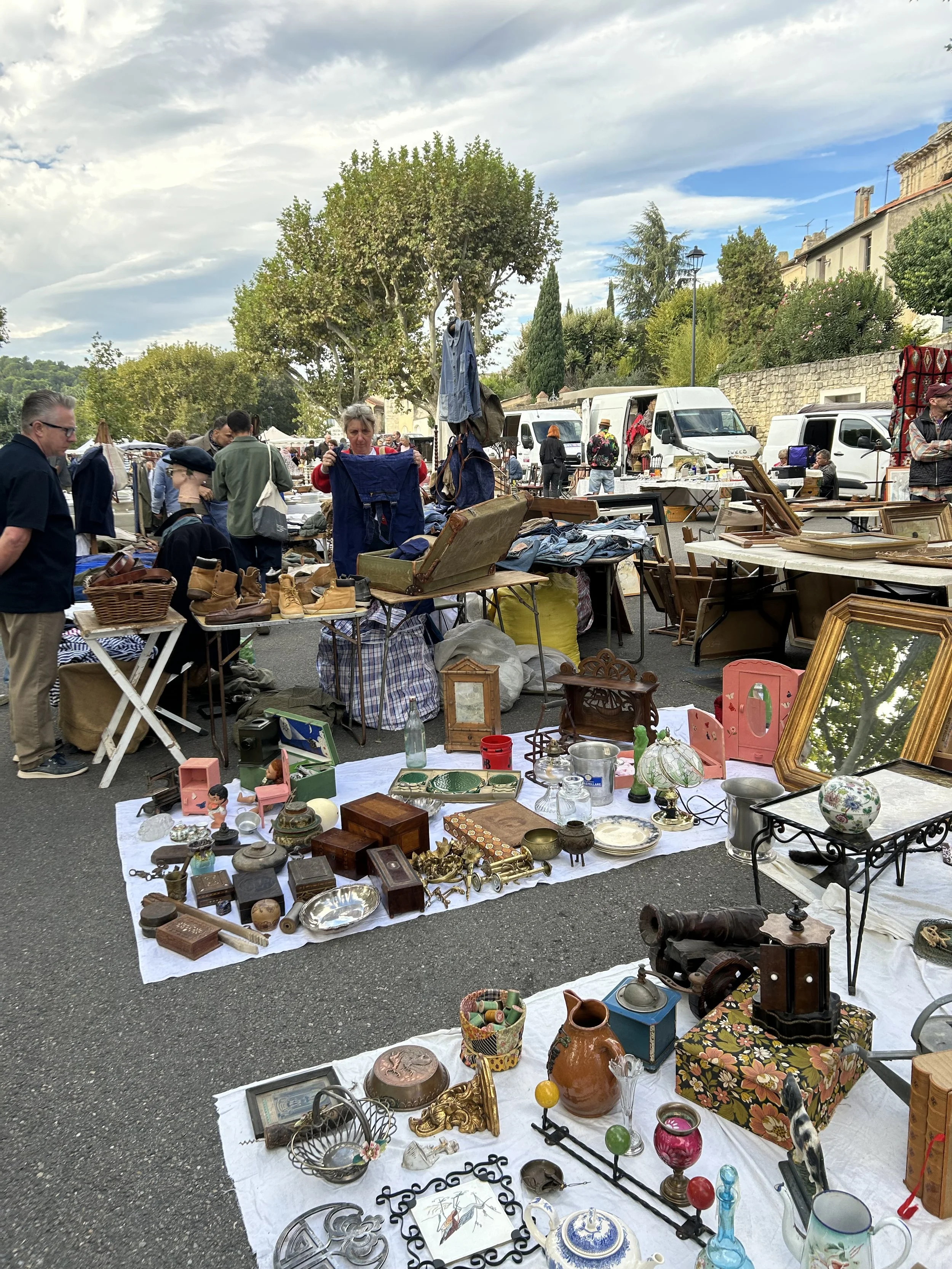 Outdoor flea market with various used items for sale including ceramics, picture frames, and decorative objects, with people browsing and vendor trucks in the background, in France