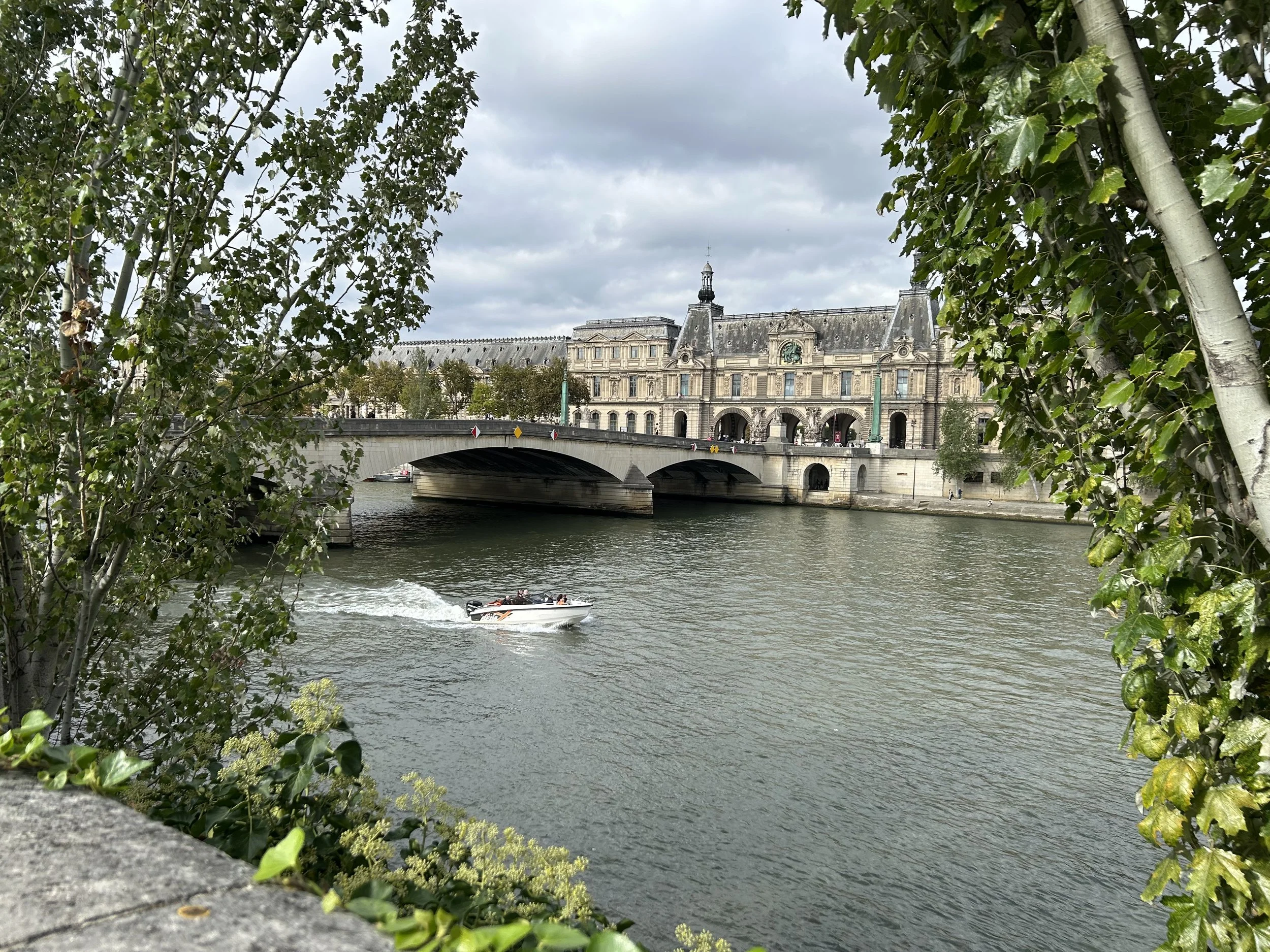 View of a historic building with ornate architecture across the Seine river, with a bridge connecting both sides. A small motorboat is on the water, and there are trees framing the scene, under a cloudy sky.