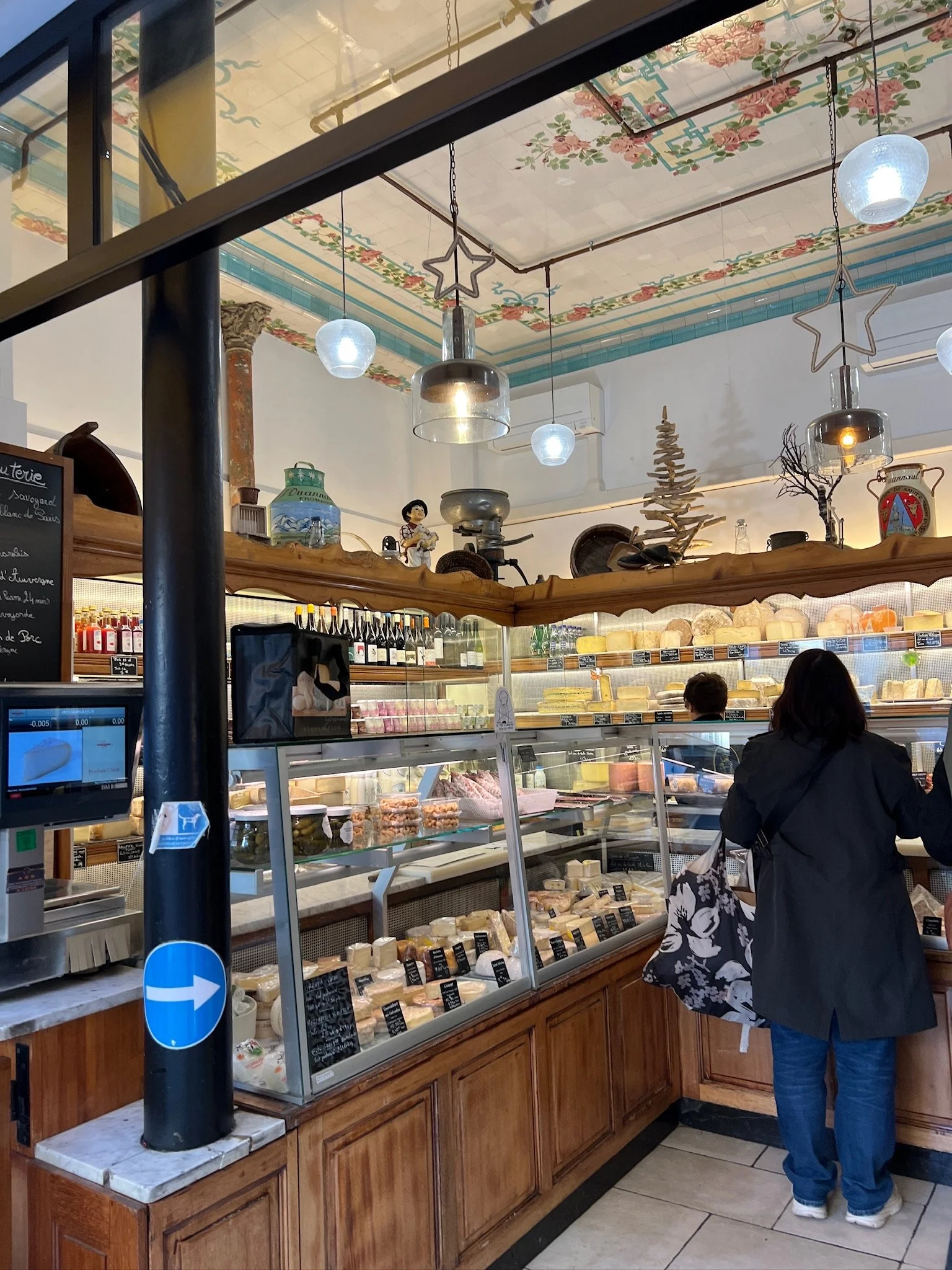 Inside a Paris cheese shop with a woman looking at cheese displays. The shop has decorative ceiling art, hanging lights, and shelves with wine bottles and vintage items.