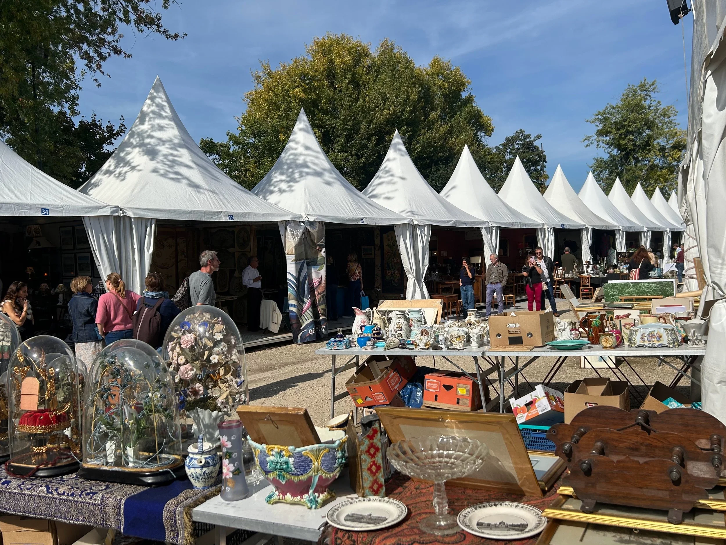 Scene at a Paris flea market with white tents, shoppers, and tables displaying various antiques and collectibles, including glassware, ceramics, and decorative objects.