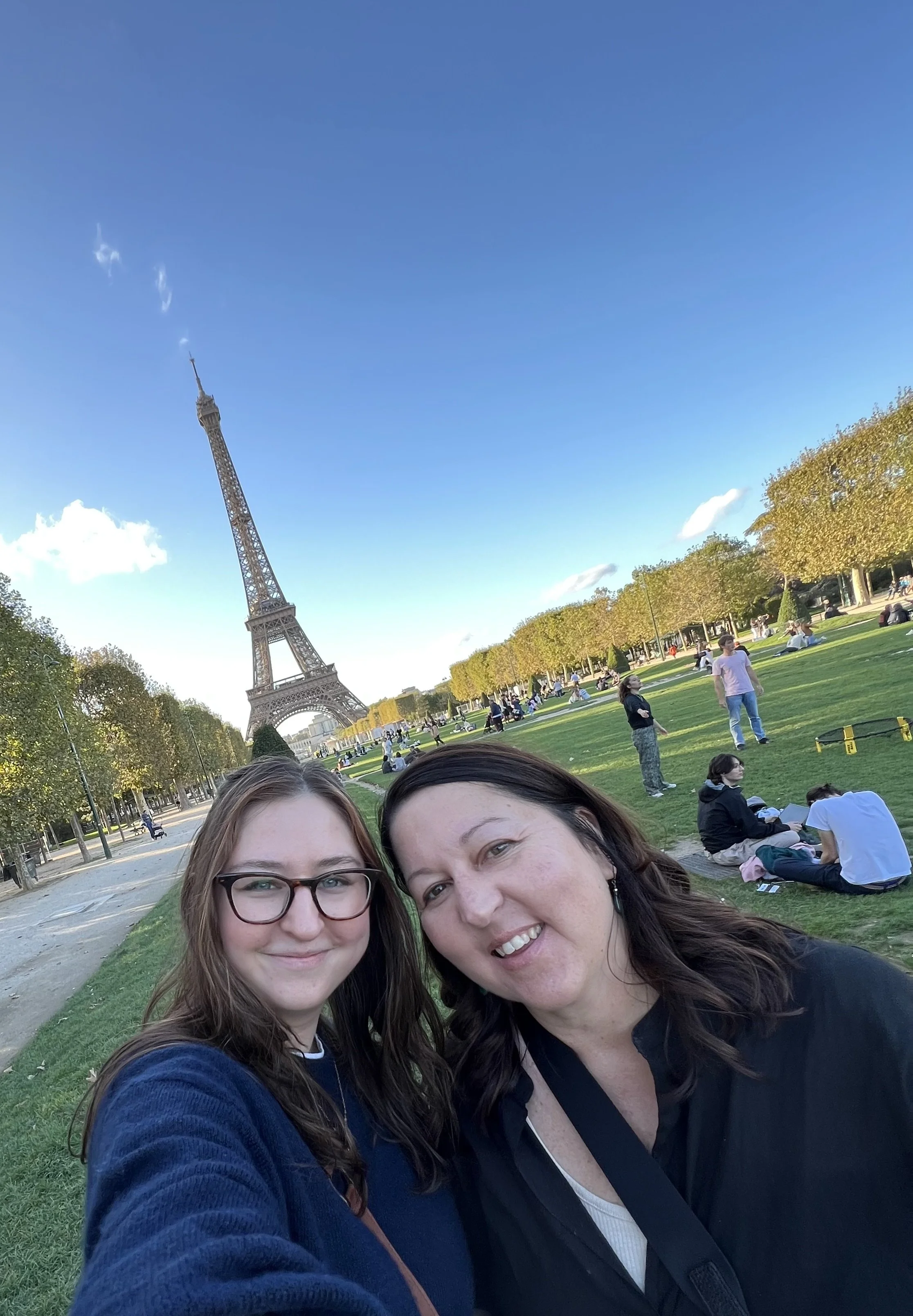 Two women take a selfie in front of the Eiffel Tower in Paris, with people sitting on the grass and walking around in the background on a clear day.