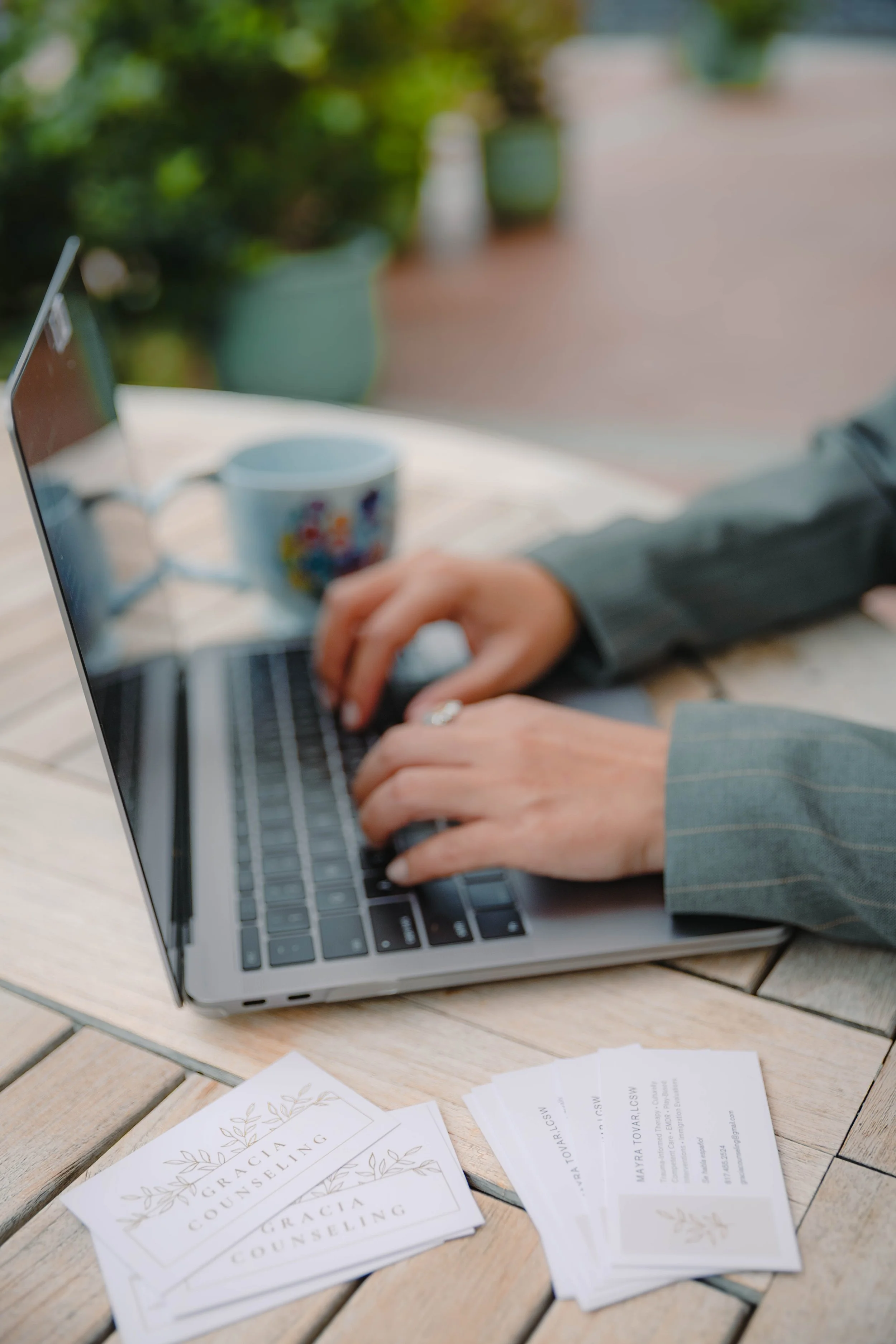Person using laptop outdoors, with business cards and a coffee mug on a wooden table.
