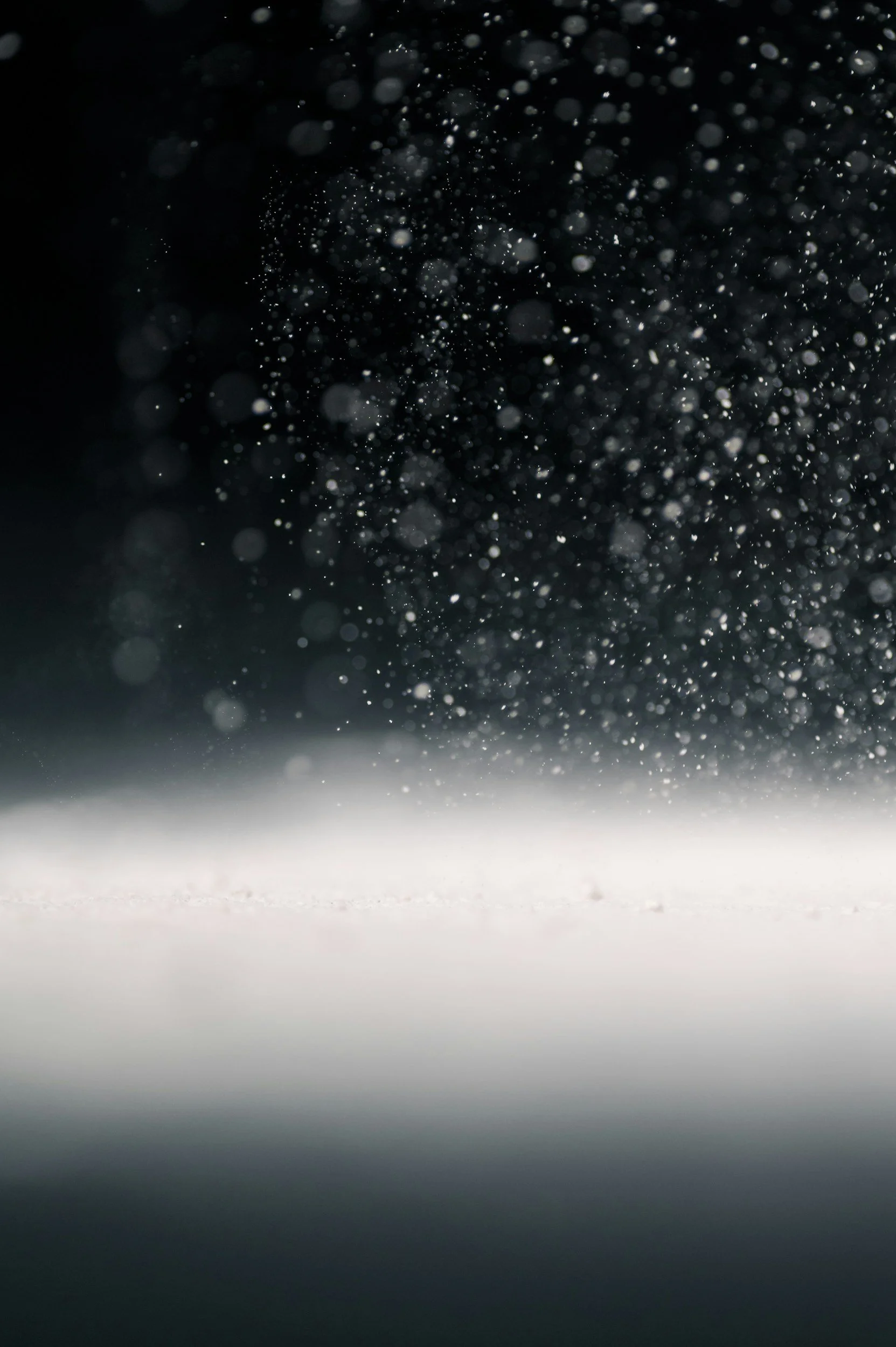 Close-up of airborne dust particles or snowflakes against a dark background with a blurred white surface below.