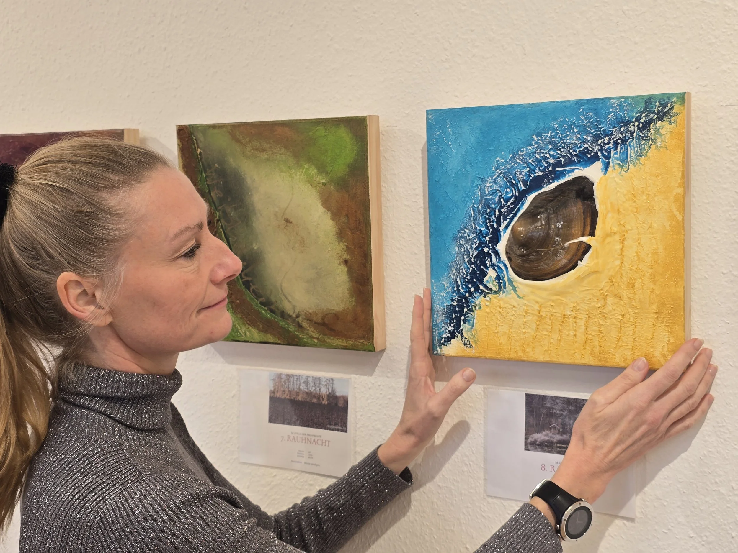 Die Künstlerin Stefanie Rau hängt in ihrer Ausstellung ein Bild aus der Serie "Rauhnächte" auf, es ist Blau und Gelb und enthält in der Mitte eine aufgesetzte Muschel. 
