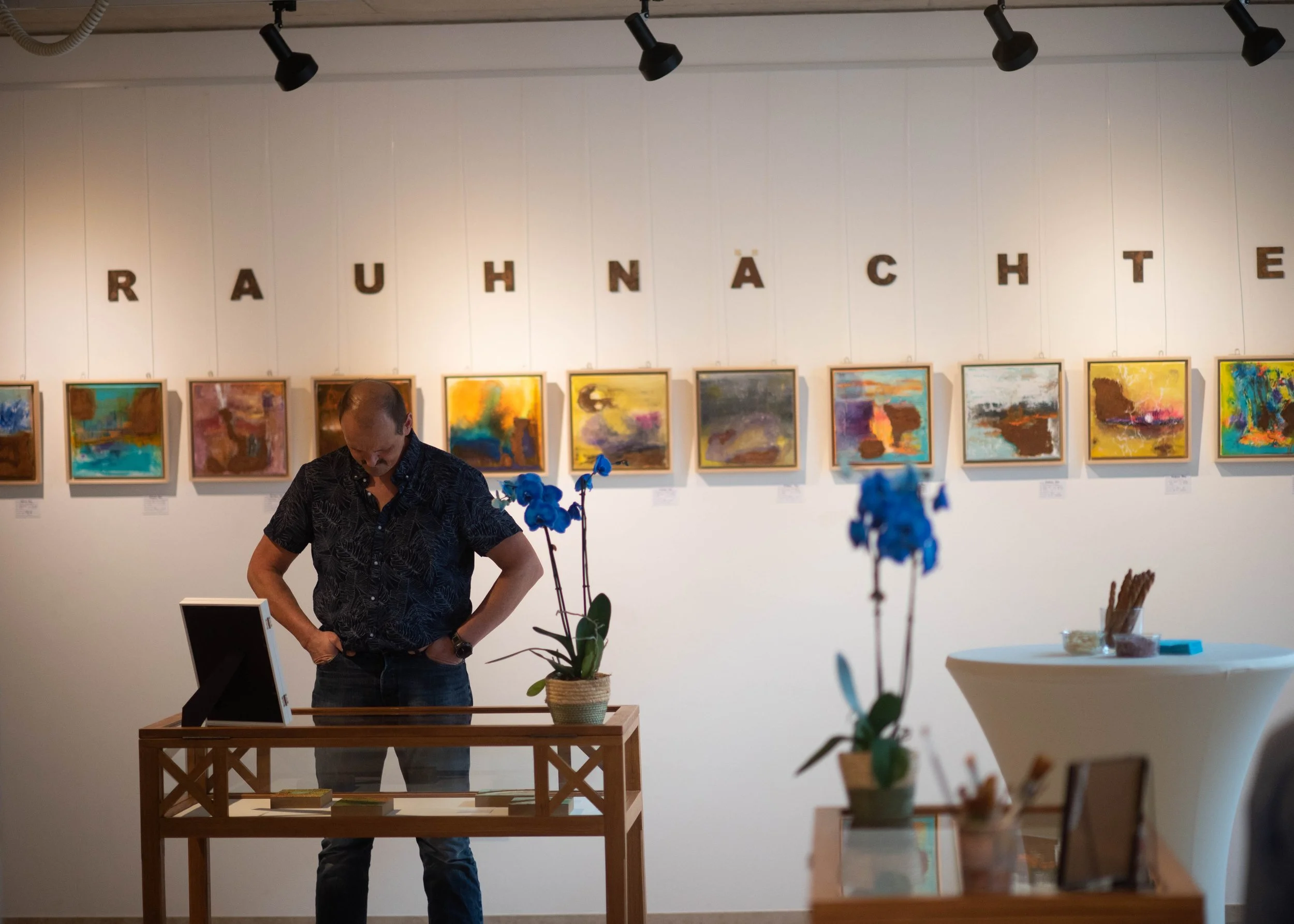Abstrakte Bilder aus der Serie "Rauhnächte 2023", ein Besucher der Ausstellung steht hinter einem Tisch und betrachtet darin ausgestellte Werke. An der Wand über den Bildern ist in rostigen Lettern das Wort "RAUHNÄCHTE" geschrieben.  