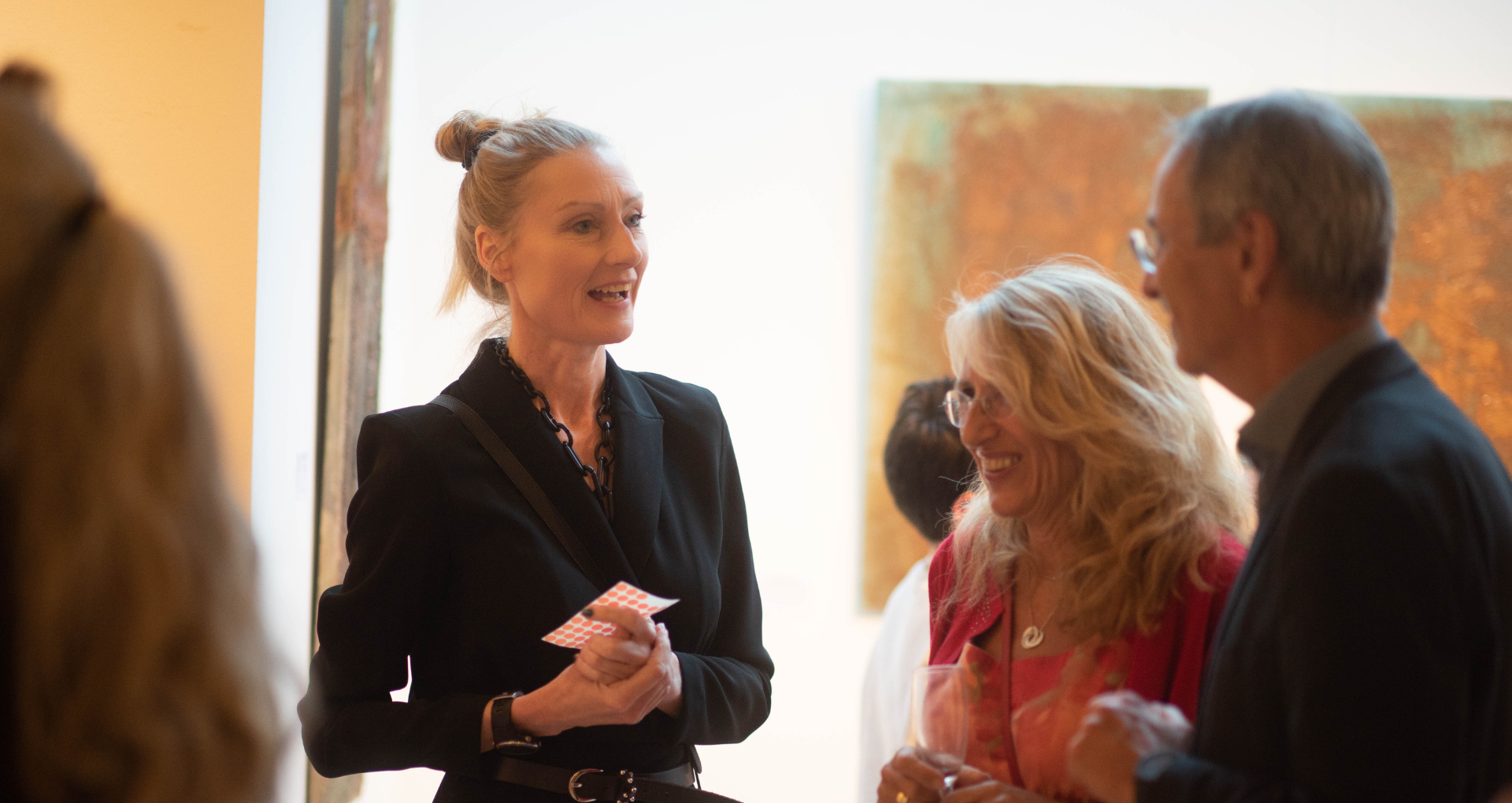 Drei Menschen beim Gespräch auf einer Kunstveranstaltung, eine Frau spricht, während die anderen beiden zuhören, im Hintergrund eine abstrakte Gemälde.