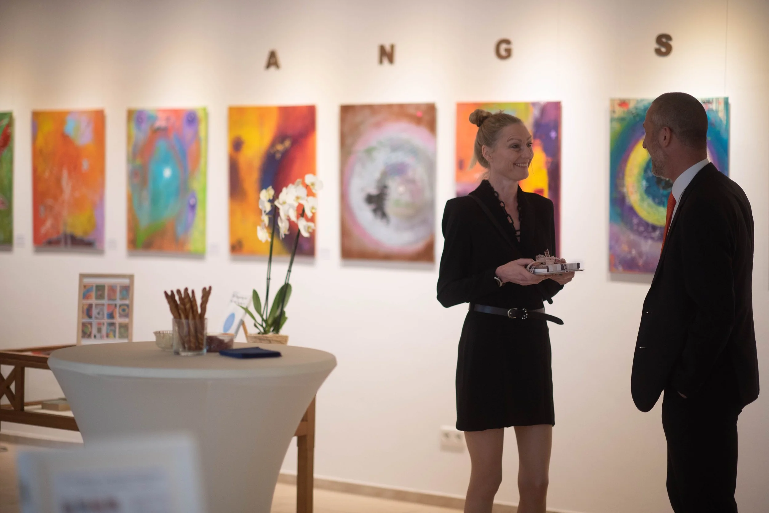 Die Künstlerin Stefanie Rau im Gespräch mit einem Gast der Vernissage im Schulmuseum Ichenhausen, im Hintergrund einige Abstrakte Werke aus ihrer ausgestellten Serie "die zwölf Ängste des menschlichen Organismus"