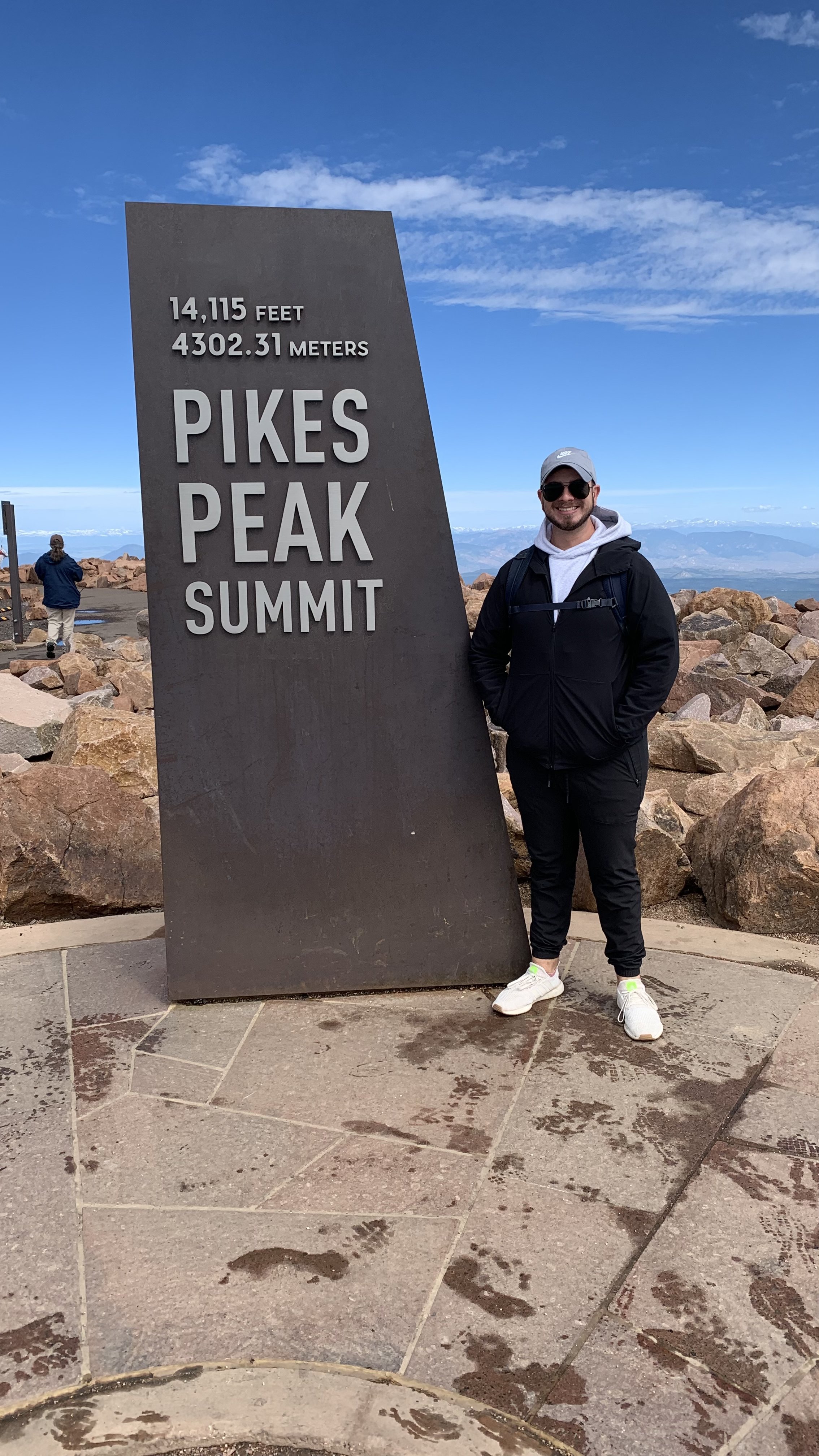 A man standing next to a sign at Pikes Peak summit, which displays its elevation of 14,115 feet and 4302.31 meters, with a mountain landscape in the background.