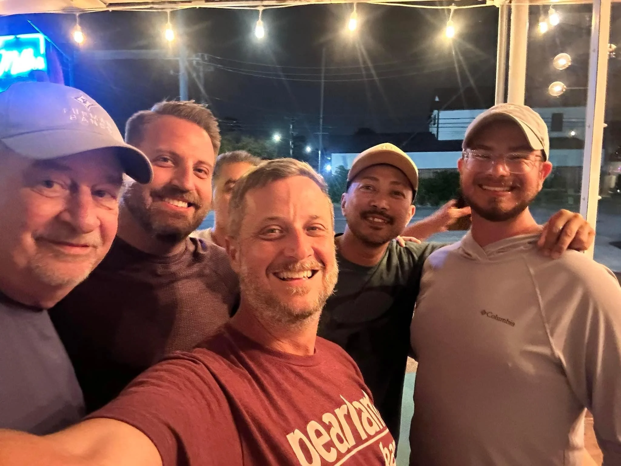 Group of six men smiling and posing for a photo at night inside a restaurant or bar with warm lighting.